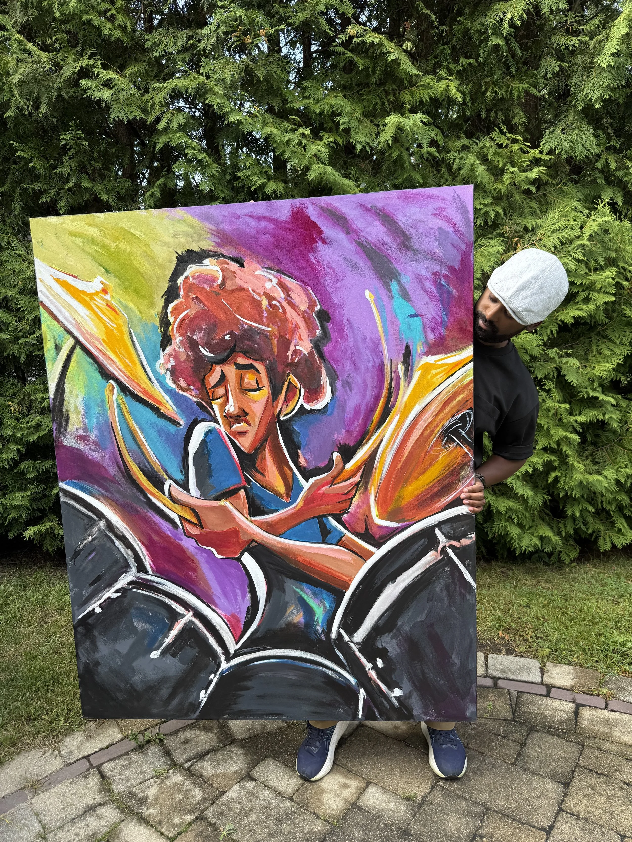 A person standing outdoors behind a large, colorful painting featuring a woman with curly hair, riding a motorcycle. The background includes green foliage and a stone-paved ground.