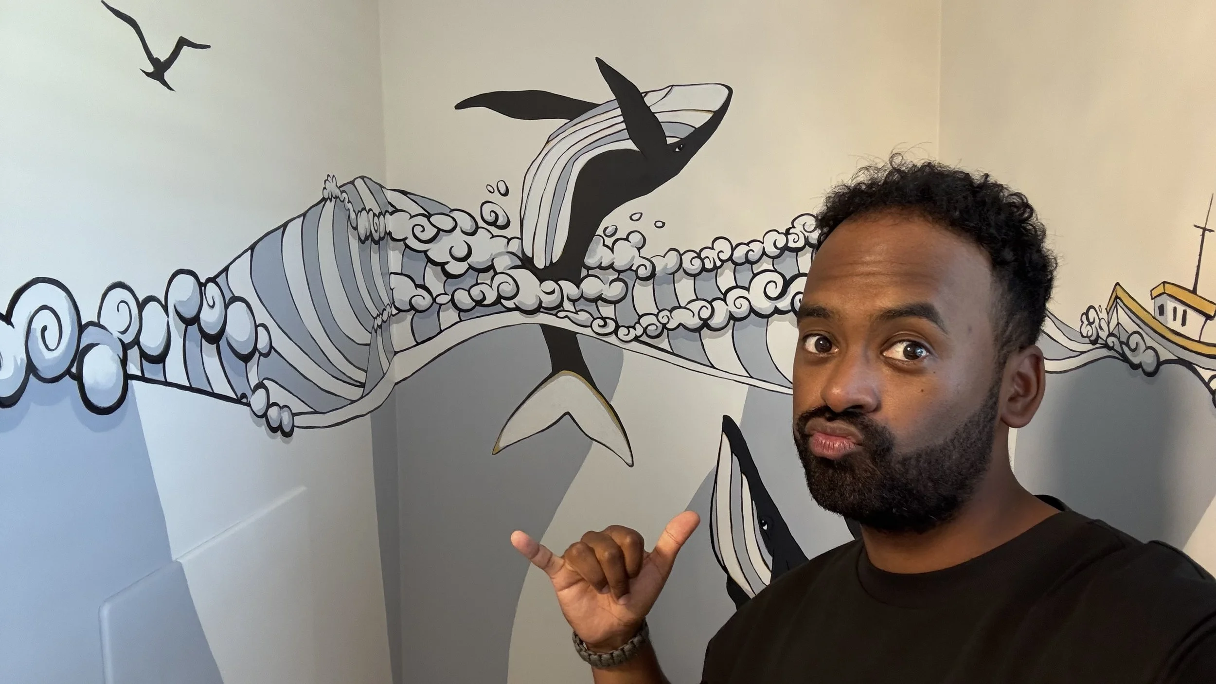 A man with curly hair and a beard taking a selfie in front of a wall art mural of a whale and birds, pointing at the mural with his right hand and making a shaka sign with his left hand.