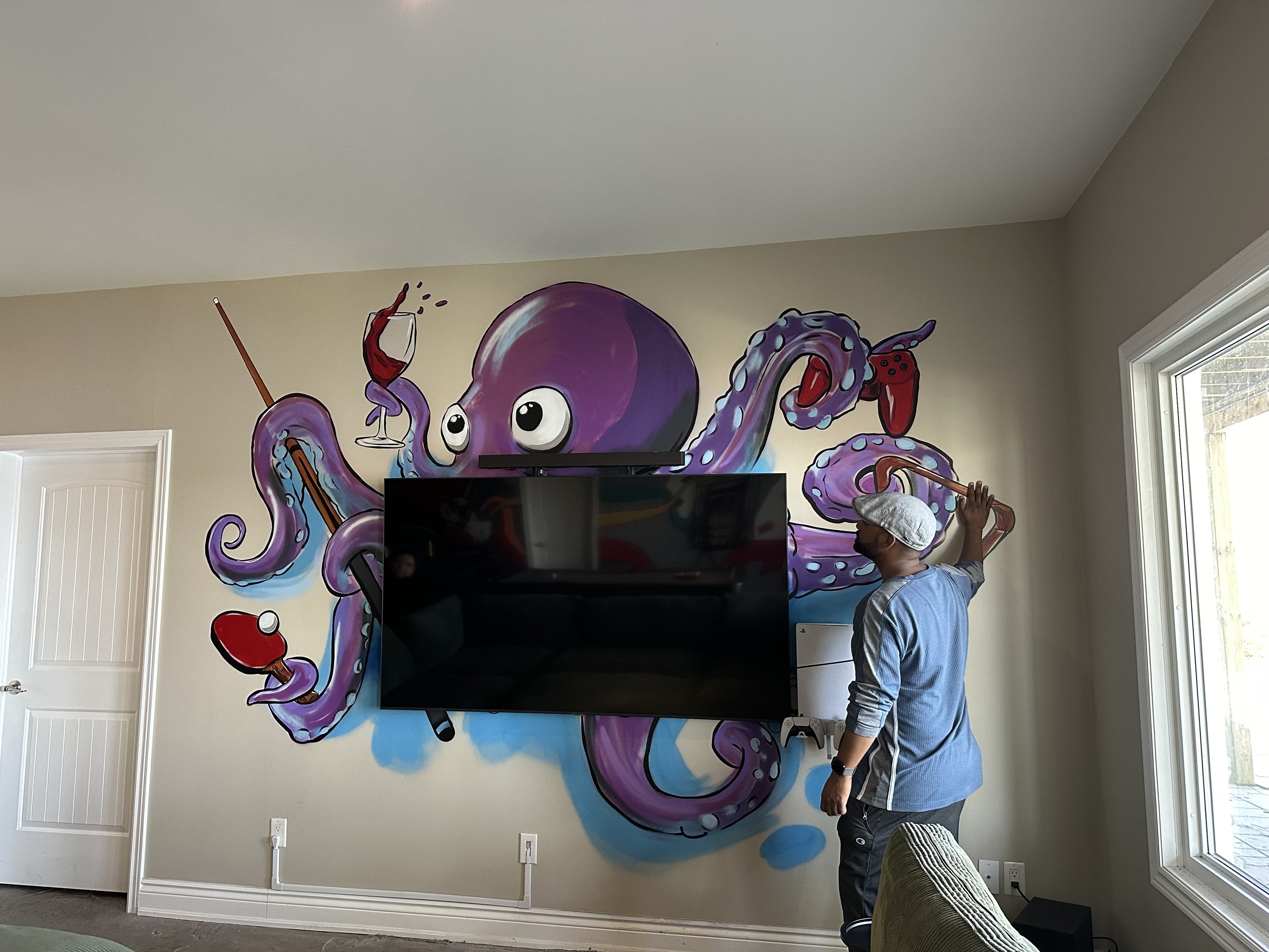 A man in a light blue jacket and gray flat cap is painting a large mural of an octopus on a beige wall. The octopus is purple with pink and light blue accents and is holding a wine glass and a paintbrush. The room has white trim, a window with sunlight, and a flat-screen TV mounted on the wall in front of the mural.