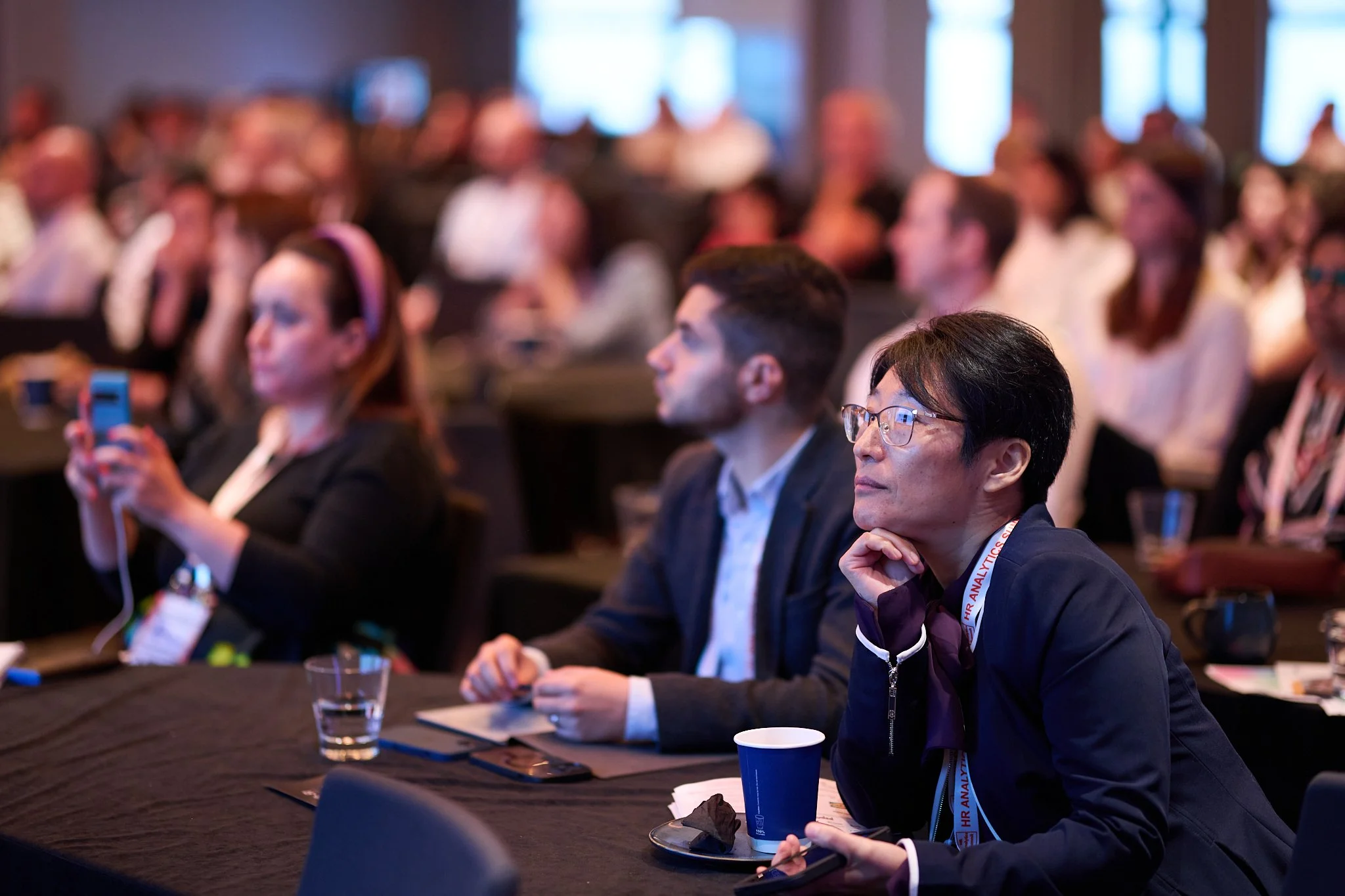 Conference attendees sitting and listening attentively, with a focus on a person in spectacles and a lanyard, holding a cup. Nearby, another attendee is using a smartphone.