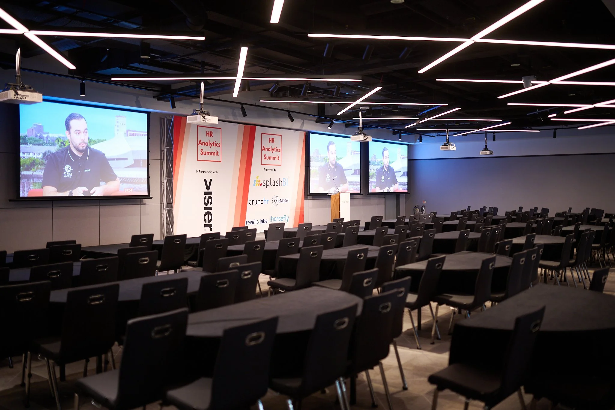 Empty conference room with round tables and chairs, large screens displaying a video, and banners for HR Analytics Summit.