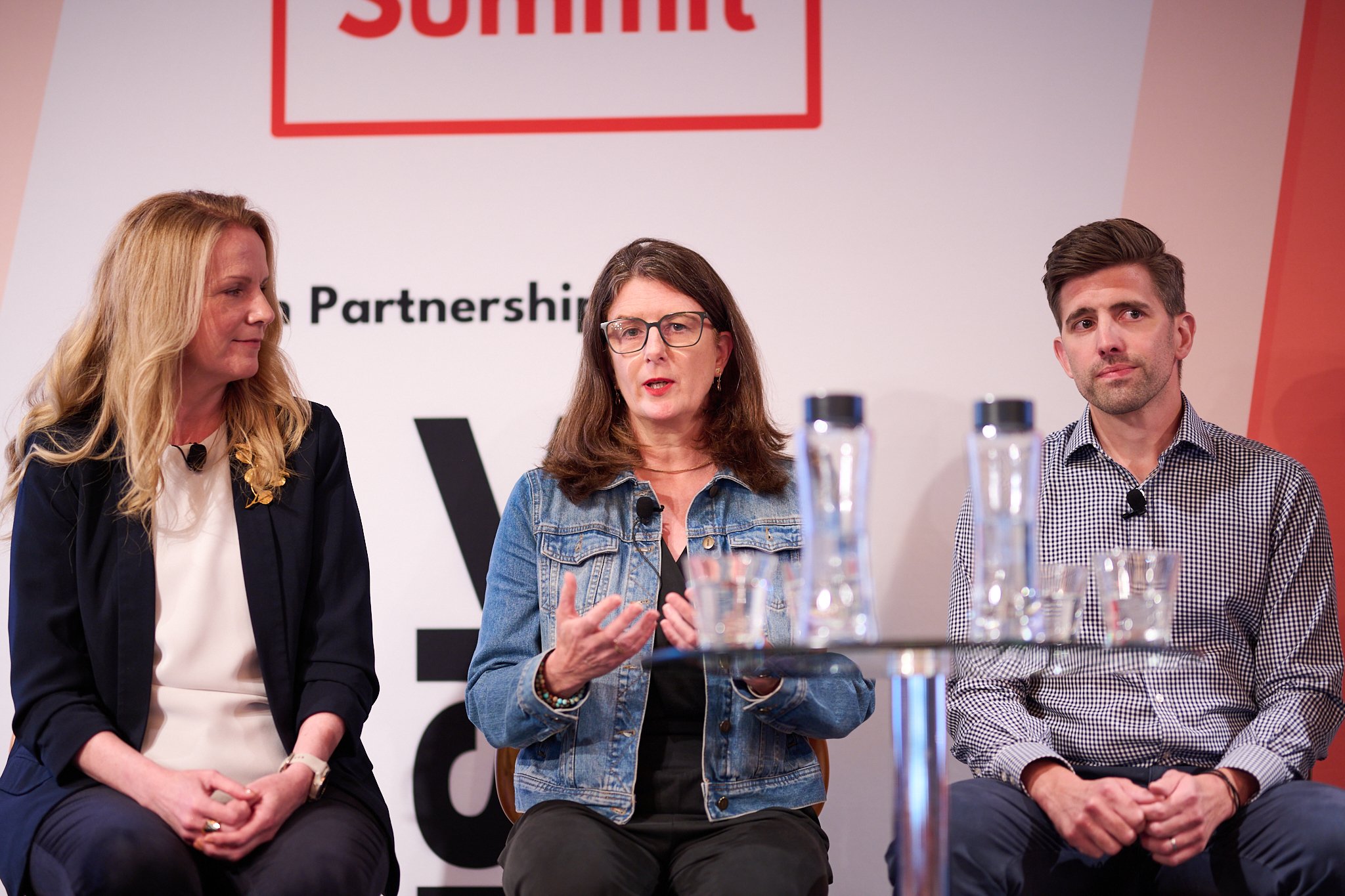 Three people sitting on a stage at a summit, one wearing a denim jacket, the others in business attire, with microphones attached.