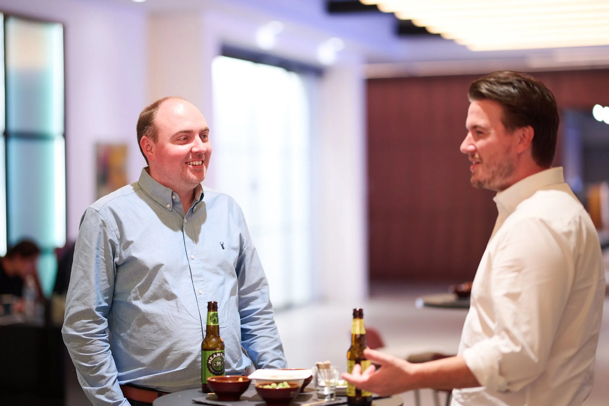 Two men in a relaxed conversation at a social event, with drinks and snacks on a table.