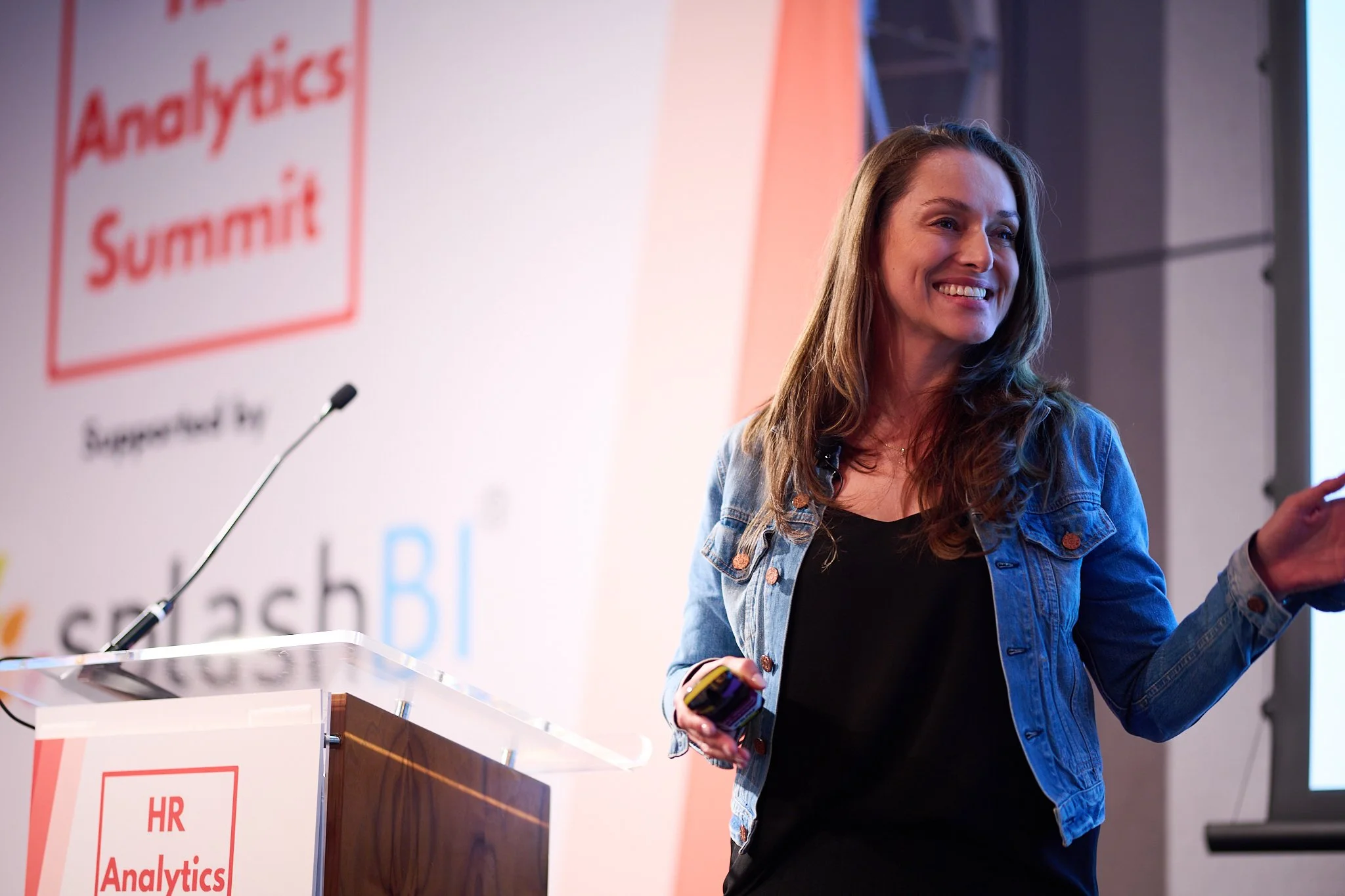 Woman speaking at HR Analytics Summit, wearing a denim jacket, with a microphone and a presentation screen in the background.