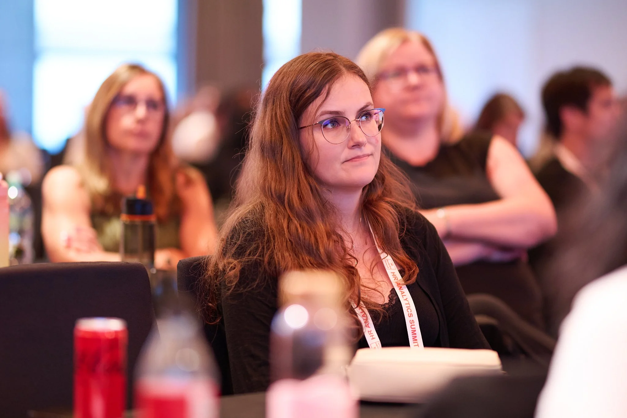 Audience in a conference setting, focusing on a woman with glasses and long hair, wearing a lanyard, with others blurred in the background.