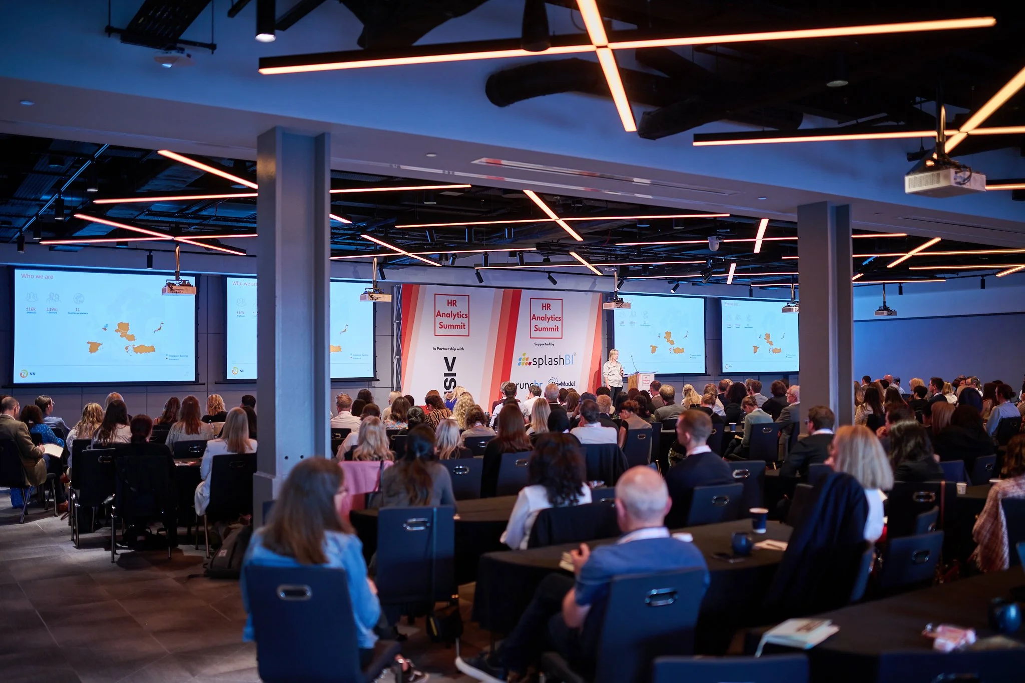 Conference attendees seated, facing a speaker at the HR Analytics Summit, with presentation screens displaying maps and text.