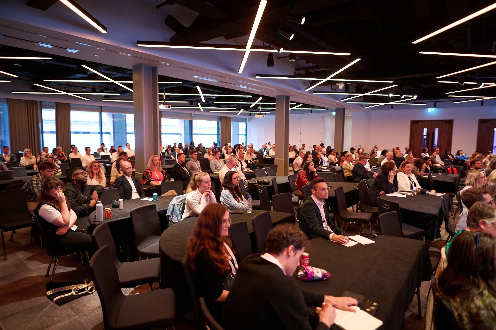 A large group of people attending a conference in a modern, well-lit meeting room with black tablecloths on the tables.