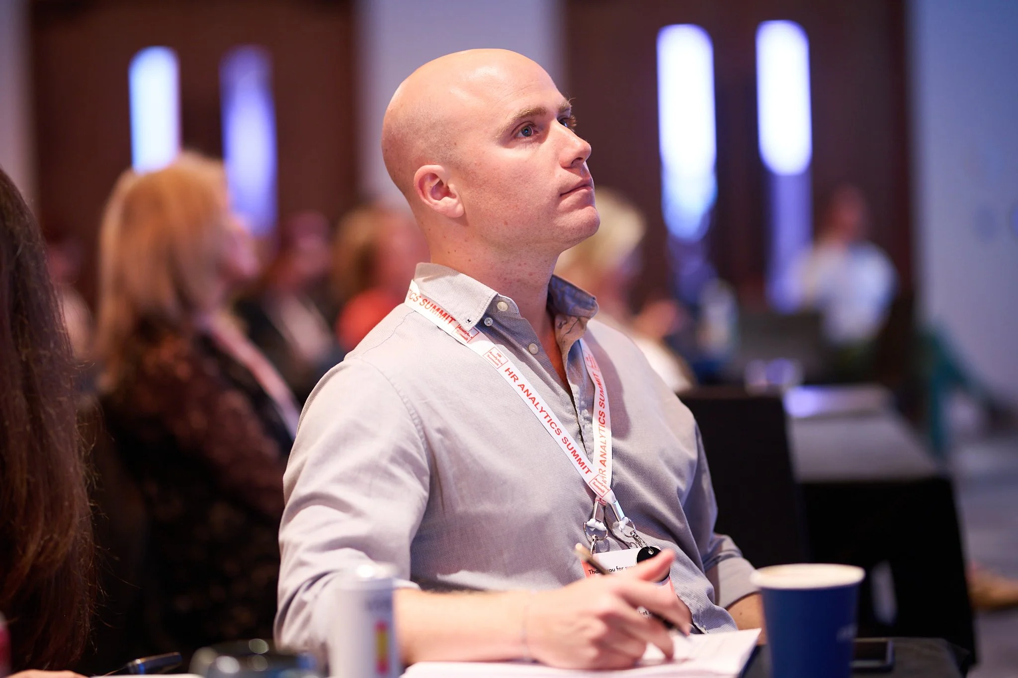 A man at a conference wearing a lanyard with "HR Analytics Summit" inscribed, attentively listening, with a pen in hand and a drink on the table.