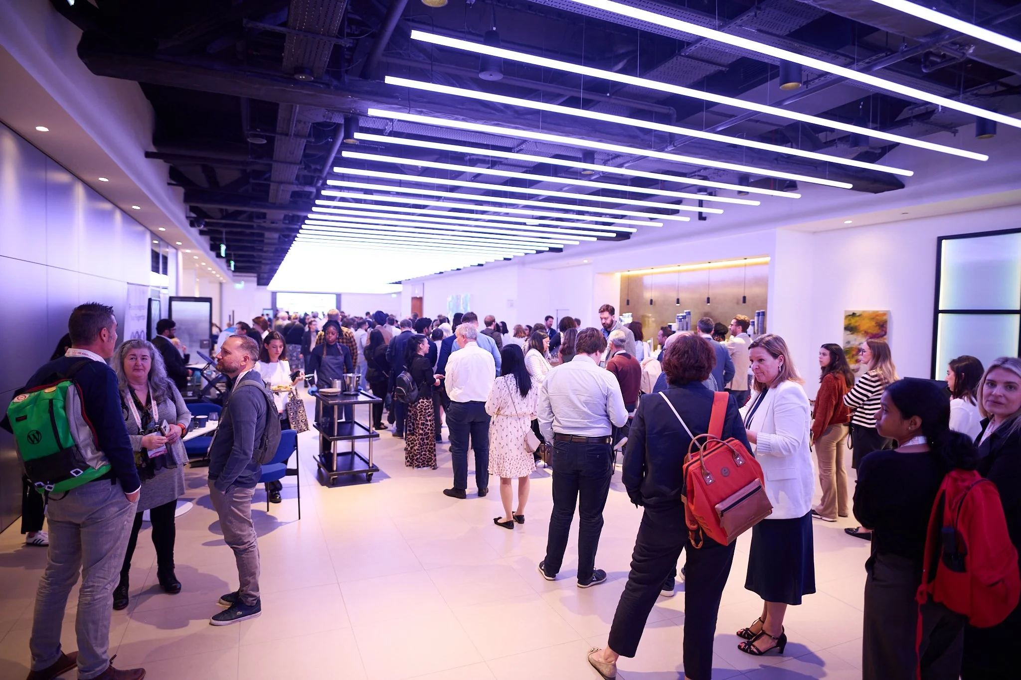 A group of people networking at an indoor event with modern lighting and decor, some wearing business attire and others casual clothes, standing and talking in a spacious room.