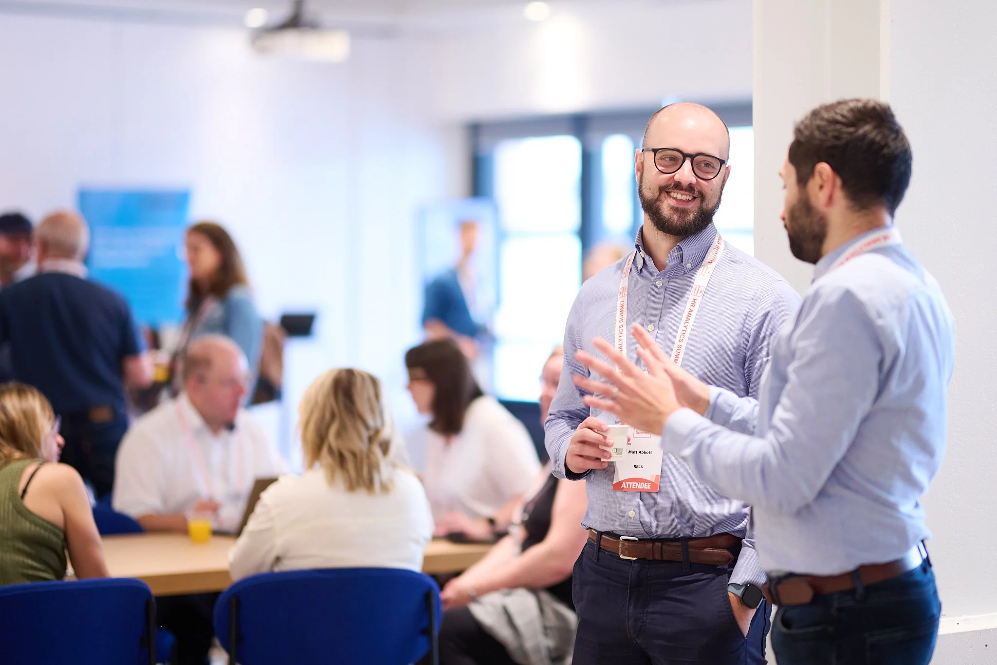 Group of people networking at a professional event, two men in foreground talking, others sitting at tables.