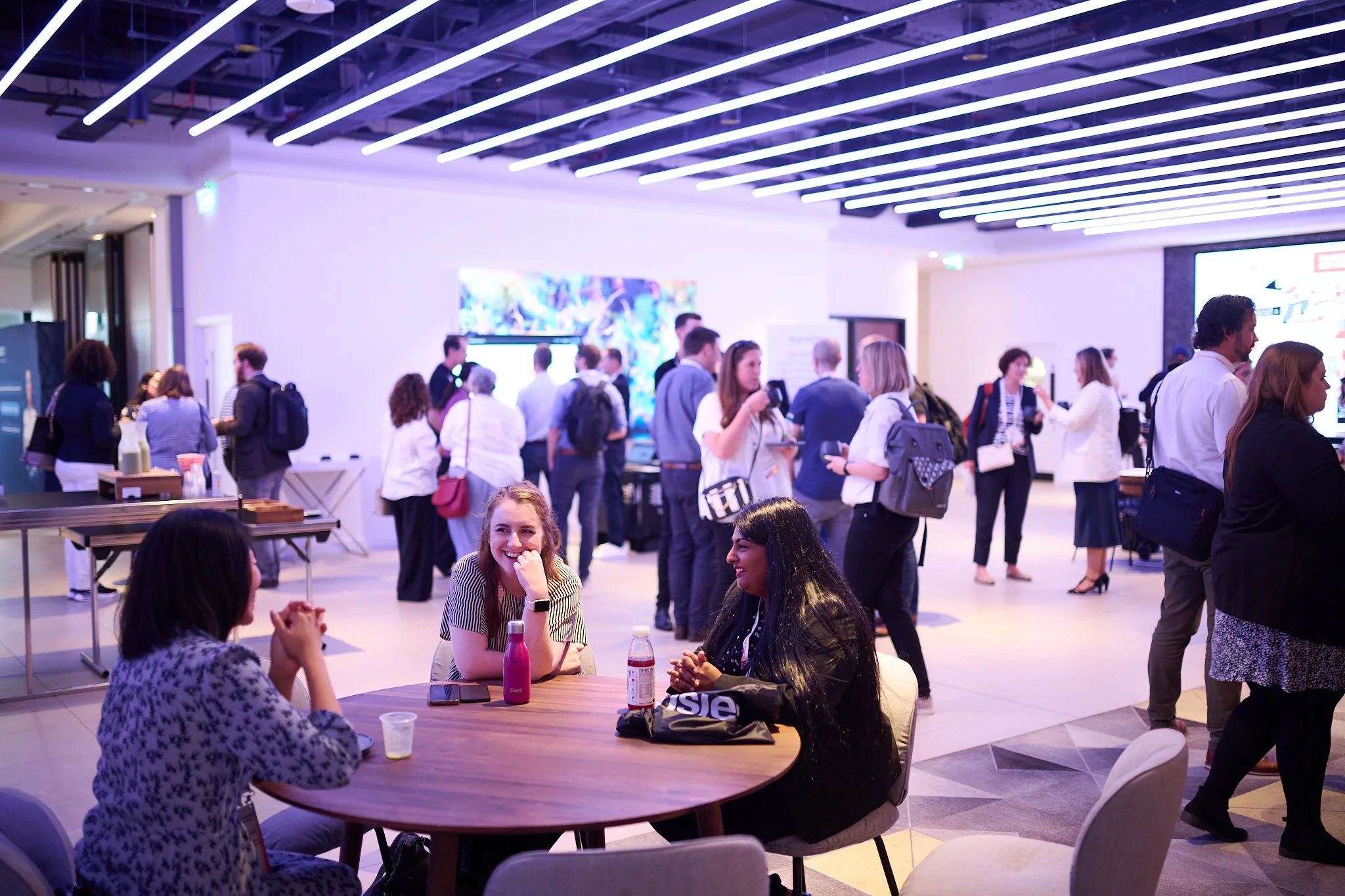 Professional networking event with people socializing in a modern conference space, featuring overhead LED lighting and a mix of standing and seated individuals.