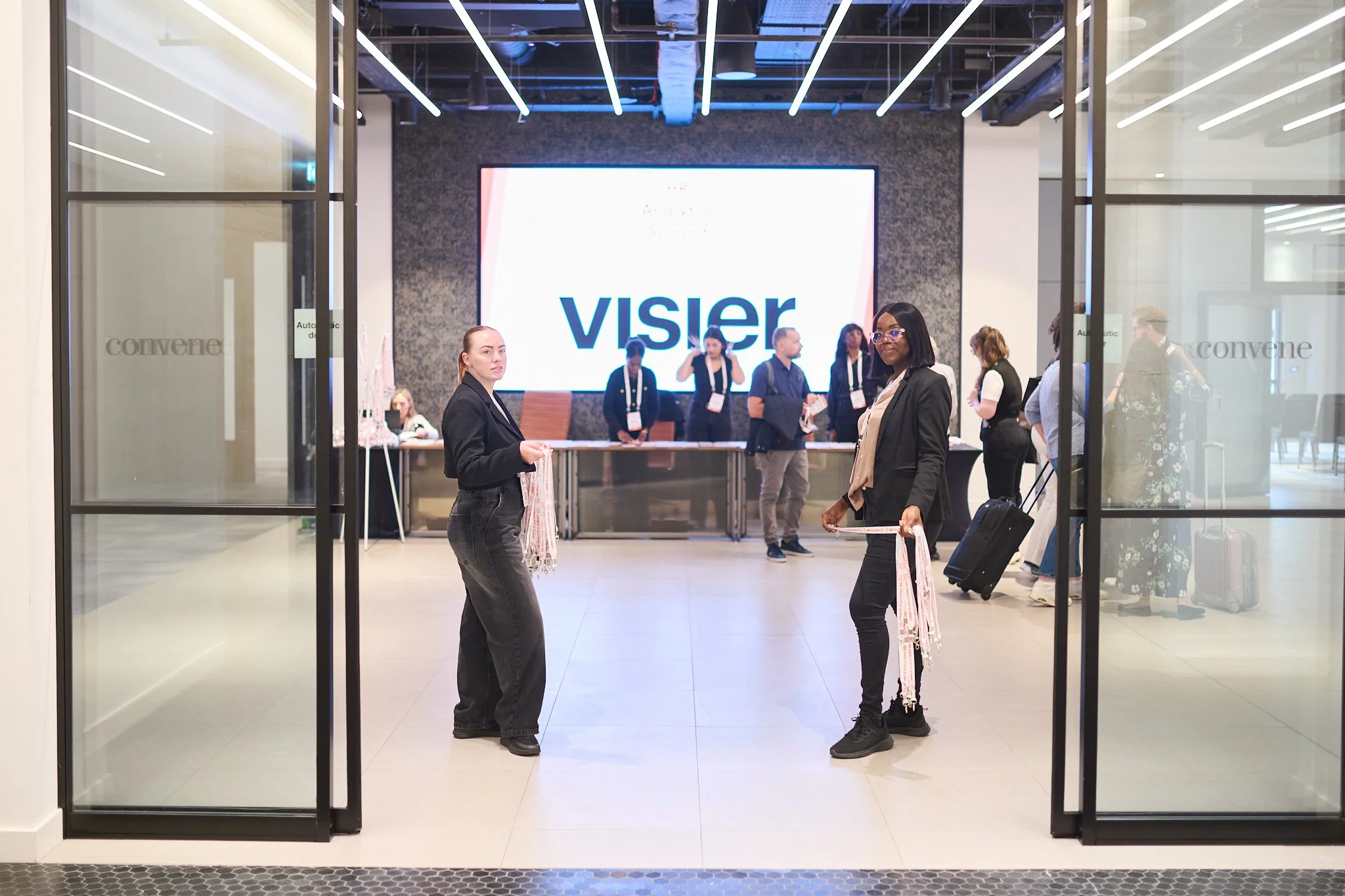 People standing at the entrance of an event venue with signage reading 'visier' in the background; attendees and staff are present, some holding lanyards.