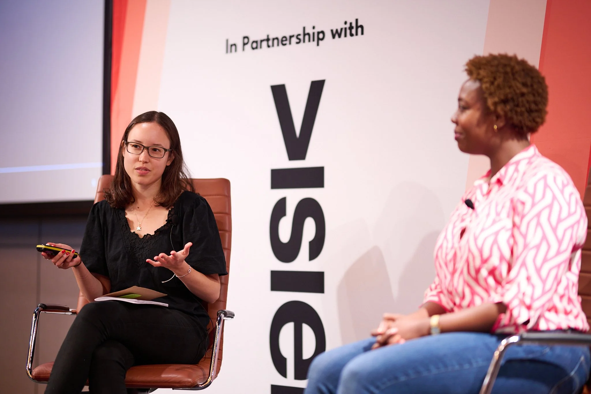 Two women engaged in a panel discussion, sitting on chairs. The woman on the left is holding a device and gesturing while speaking. A "In Partnership with..." banner is partially visible in the background.