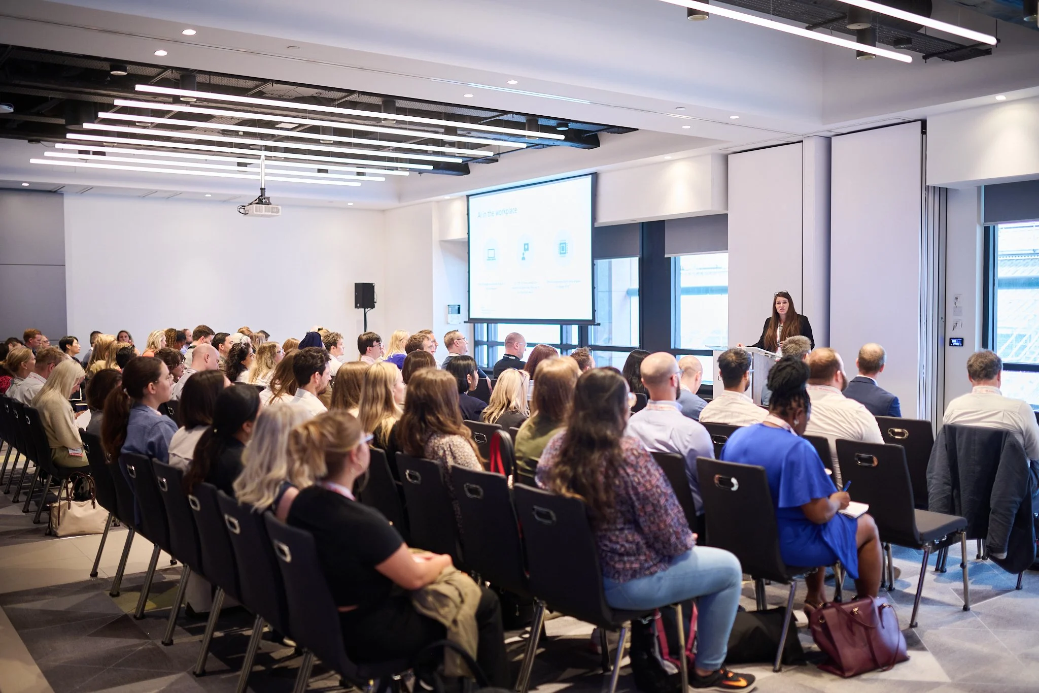 A conference room with a speaker presenting to an audience seated in rows facing a large screen.