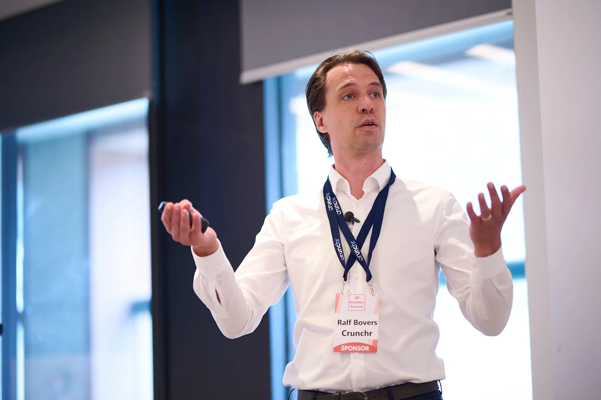 A man speaking at a conference wearing a white shirt and holding a presentation device, with a name tag labeled "Ralf Bovers, Crunchr, Sponsor."