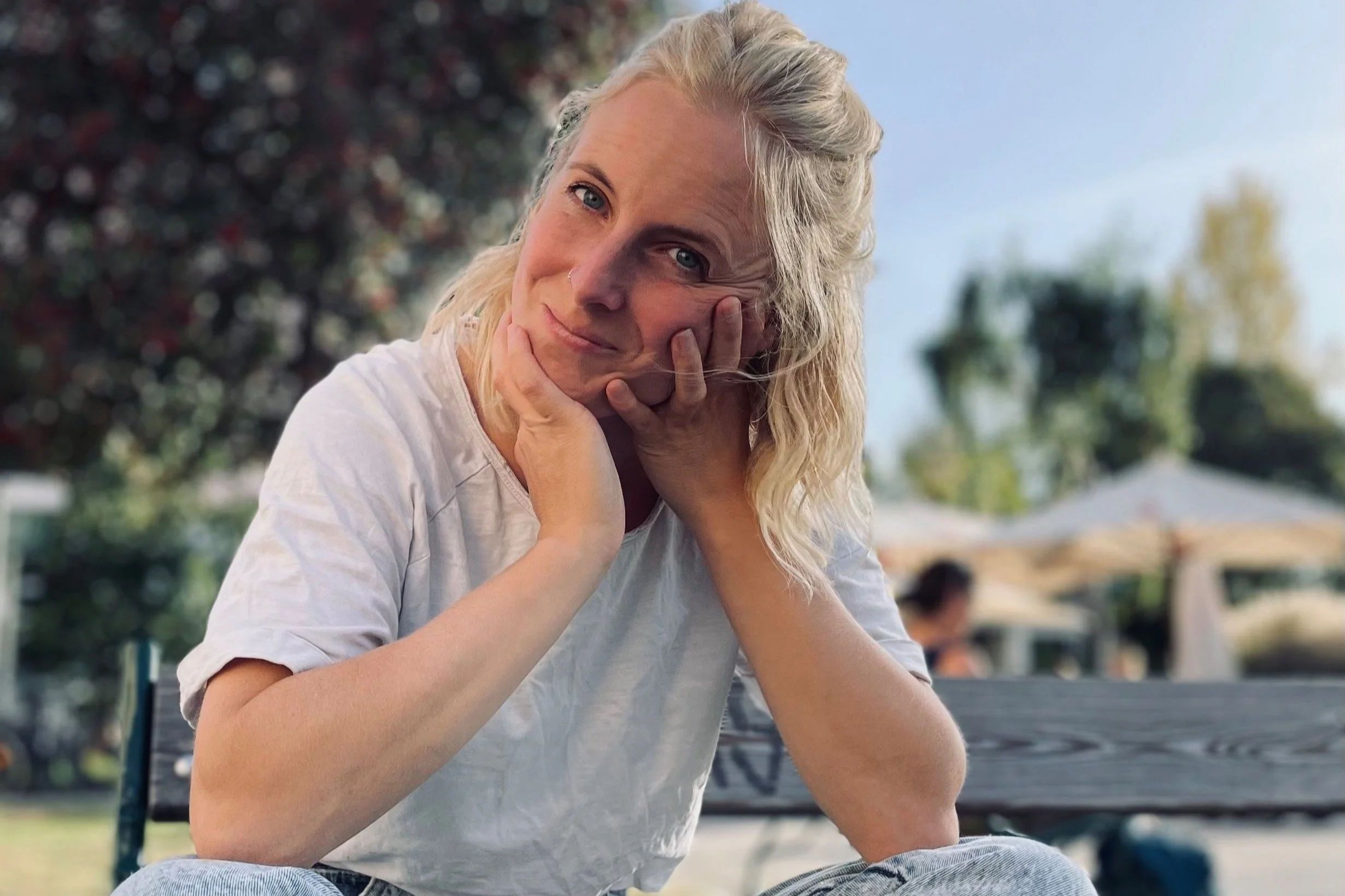 Eine junge Frau mit blondem, gewelltem Haar sitzt draußen auf einer Bank im Park, lächelt, im Hintergrund sind Bäume und der blaue Himmel sichtbar.