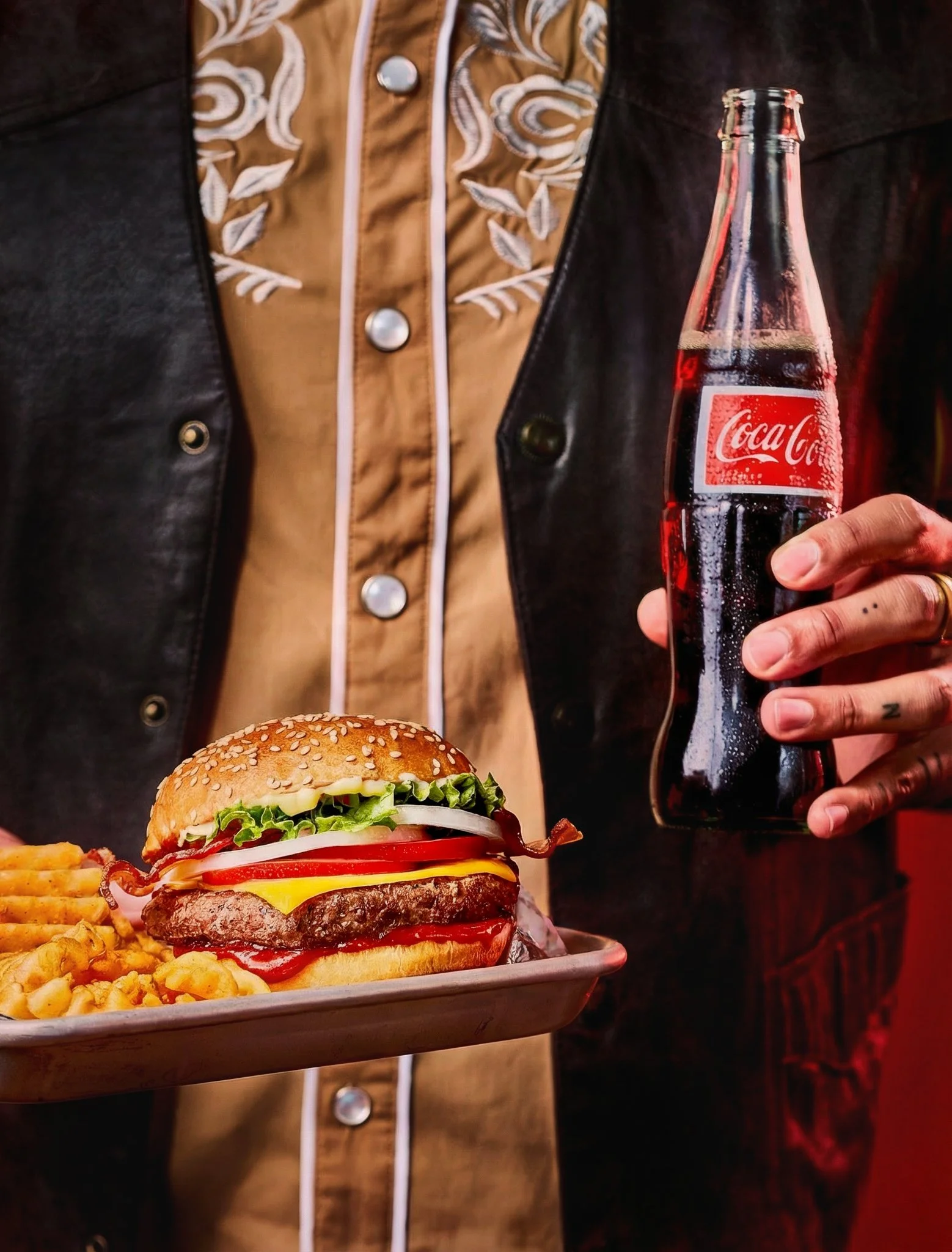 Person holding a Coca-Cola bottle and serving a cheeseburger with French fries on a tray. Styled cheeseburger with French fries and Coca-Cola on a tray, commercial food photography by Joni Schrantz and food stylist Chris Caldes in Denver, Colorado