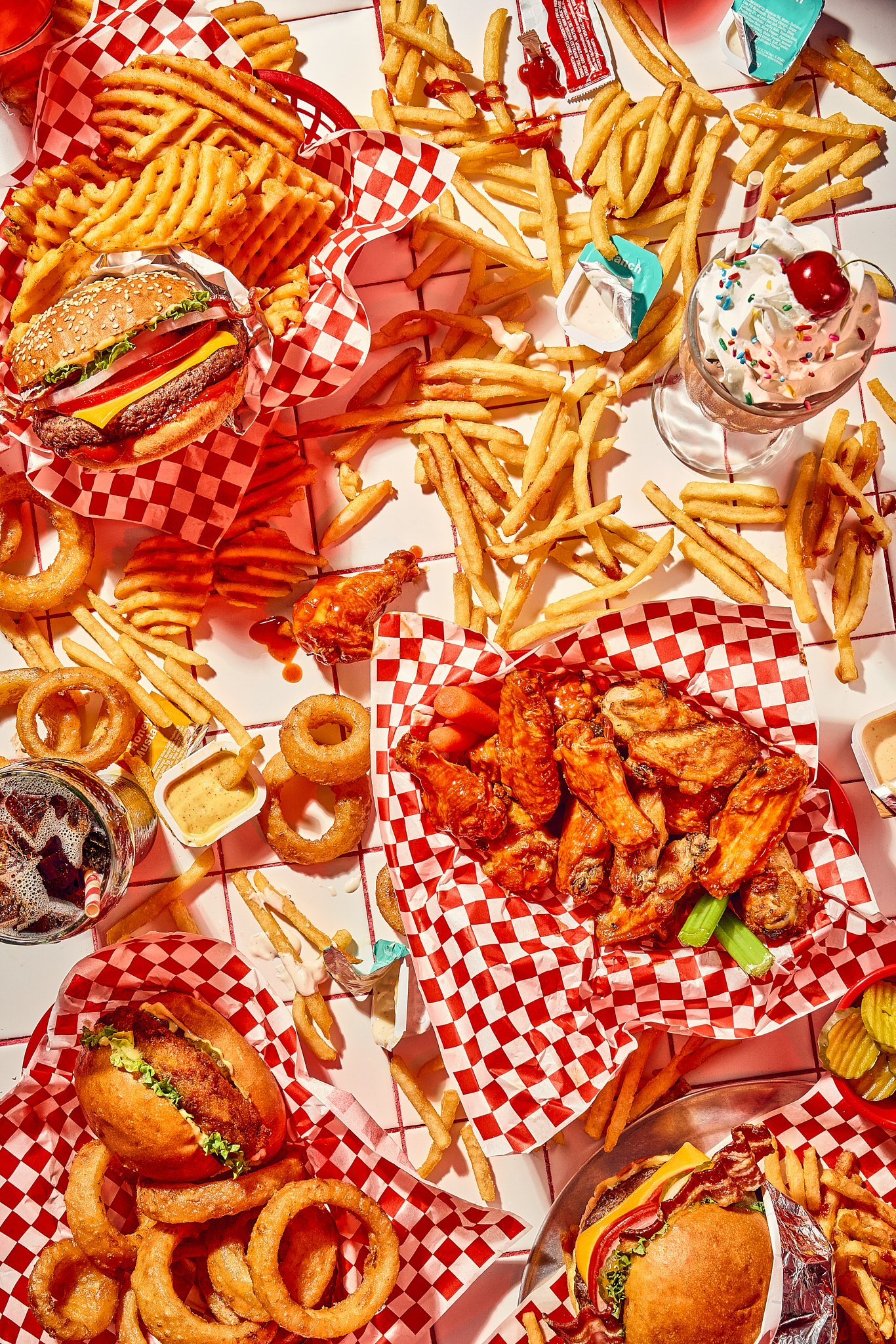 A spread of fast food including cheeseburgers, chicken wings, onion rings, French fries, and milkshakes on a table with red and white checkered paper. Professional food photography by Joni Schrantz and one of Denver's top food stylists, Chris Caldes.