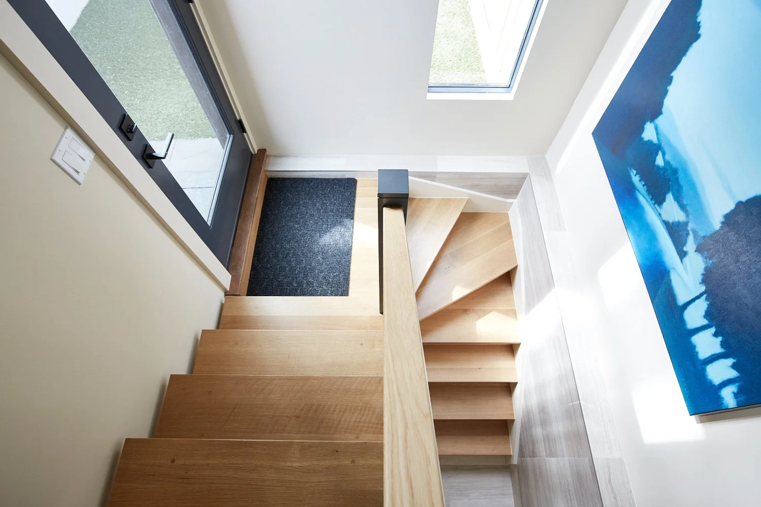 Interior view of a staircase with a wooden handrail, black doormat, a glass door with a metal frame, a window, and part of a blue landscape painting on the wall.