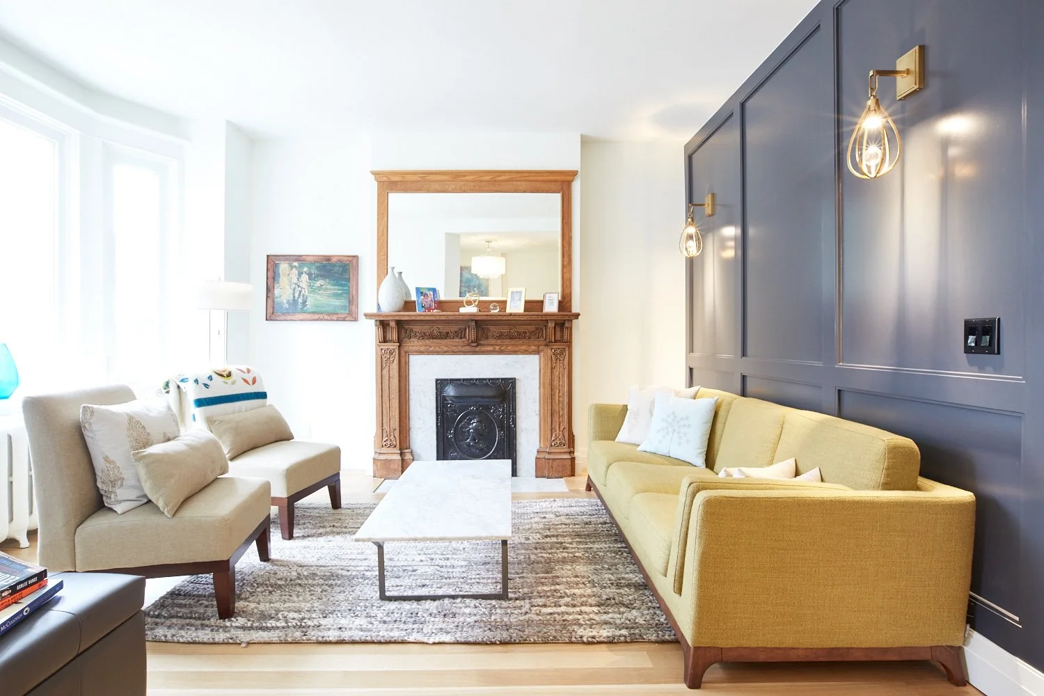 Living room with two sofas, one beige and one yellow, a fireplace with a wooden mantel, a large mirror above it, and wall-mounted lights on a dark blue accent wall.
