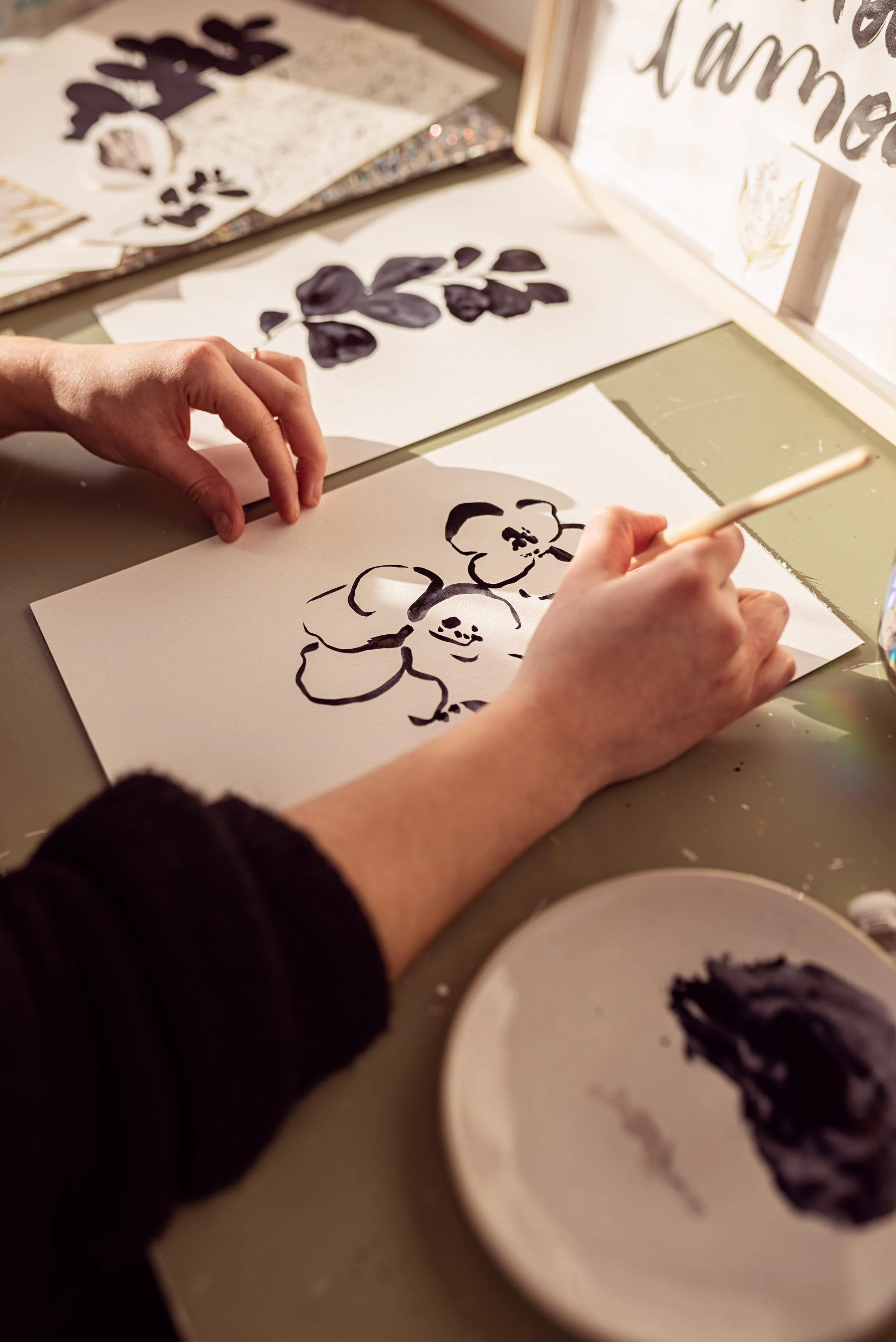 Reportage photo créateur pour la Presse. Personne en train de peindre une illustration florale en noir sur une feuille de papier blanc, avec des autres œuvres florales autour. Éclairage naturel à proximité d'une fenêtre.