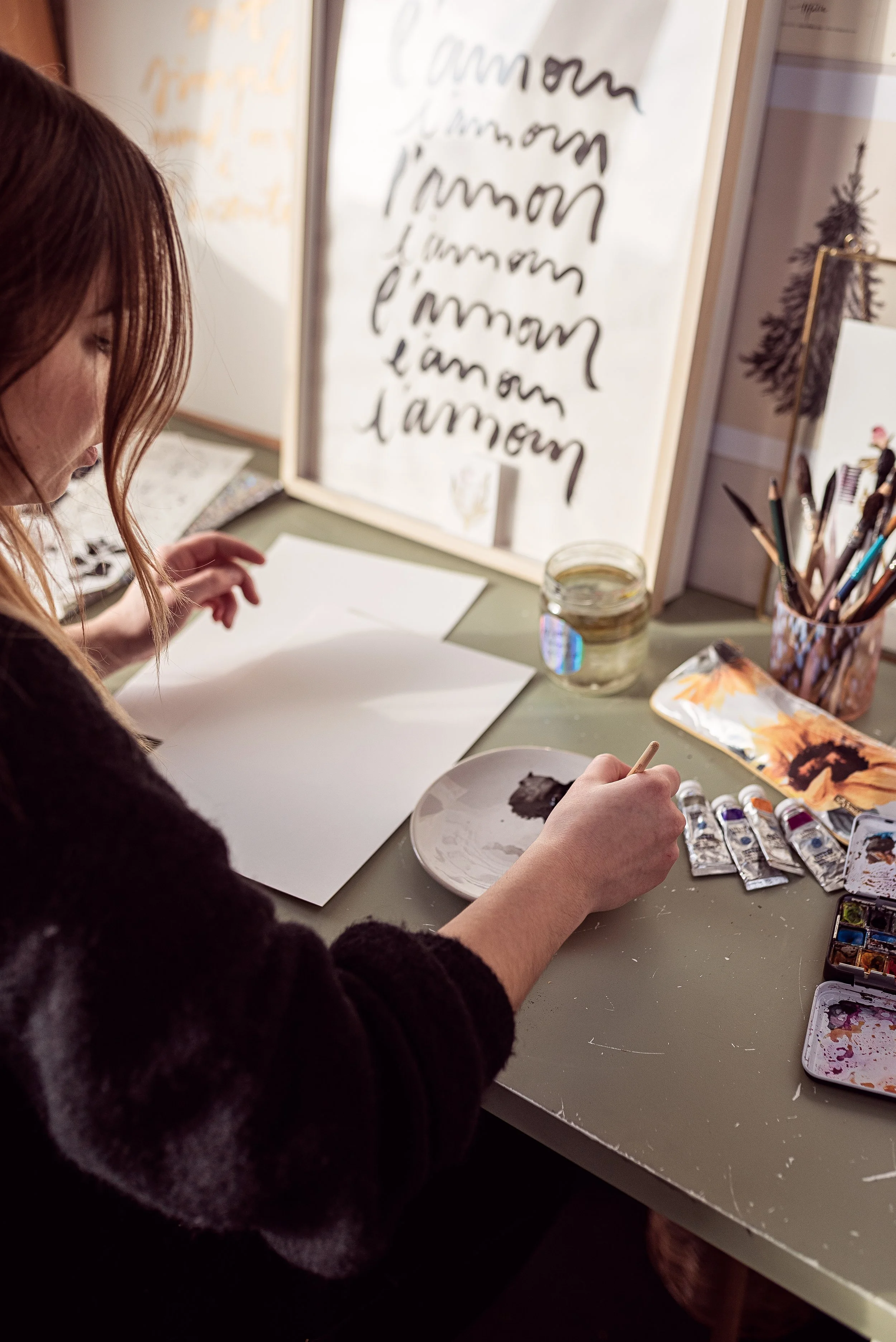 Une femme en train de peindre ou de dessiner à une table avec des tubes de peinture, un petit plat pour mélanger la peinture, un pinceau et un cahier blanc. En arrière-plan, une affiche avec l'écriture répétée du mot 'l'amour' et une petite décoratio