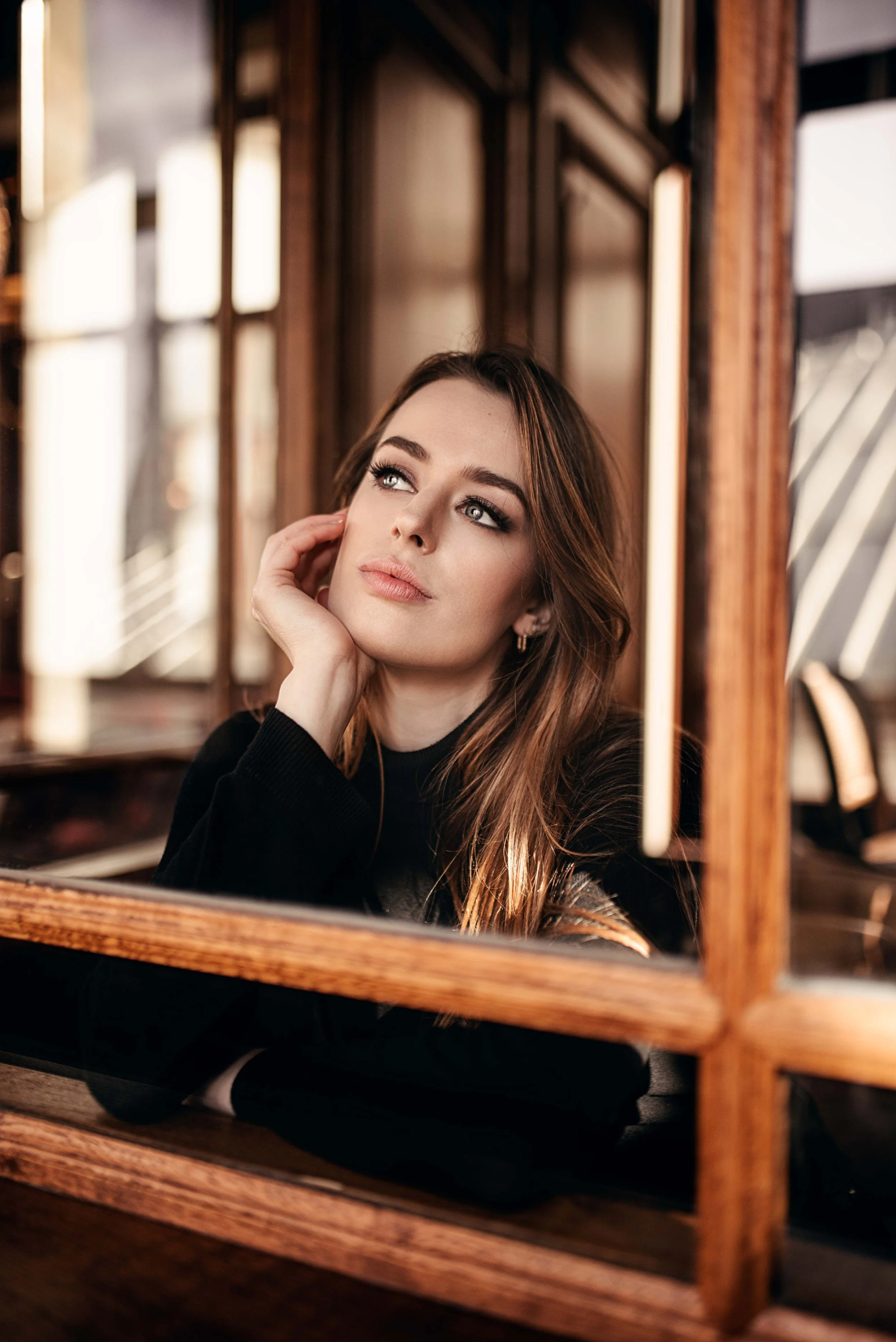Une femme aux cheveux bruns, portant un pull noir, regarde pensivement à travers une fenêtre en bois dans un café ou restaurant.