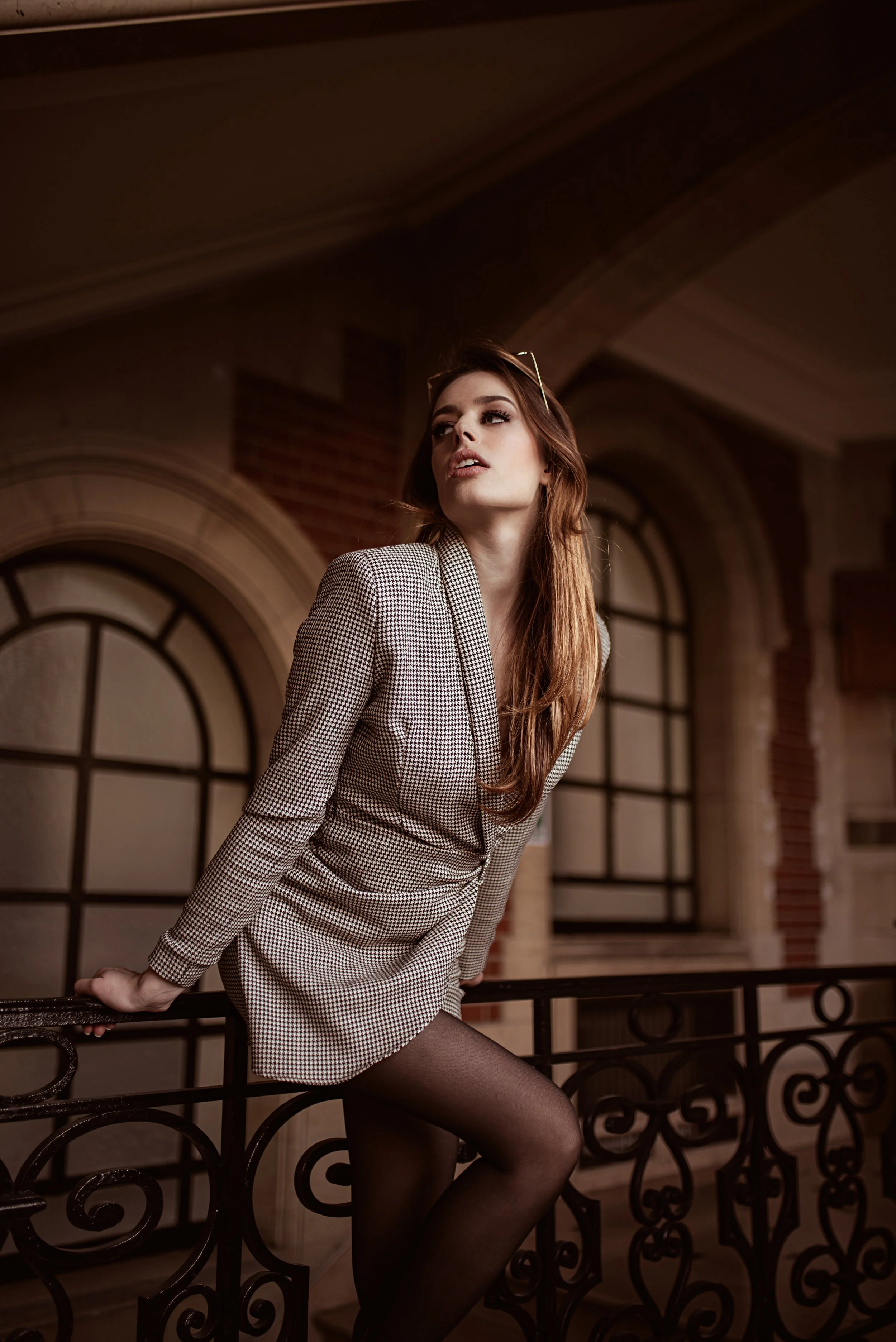 Une femme aux cheveux roux, portant une veste à motifs, pose sur un balcon en bois dans un bâtiment ancien avec de grandes fenêtres arched, dans une ambiance chaleureuse et élégante.
