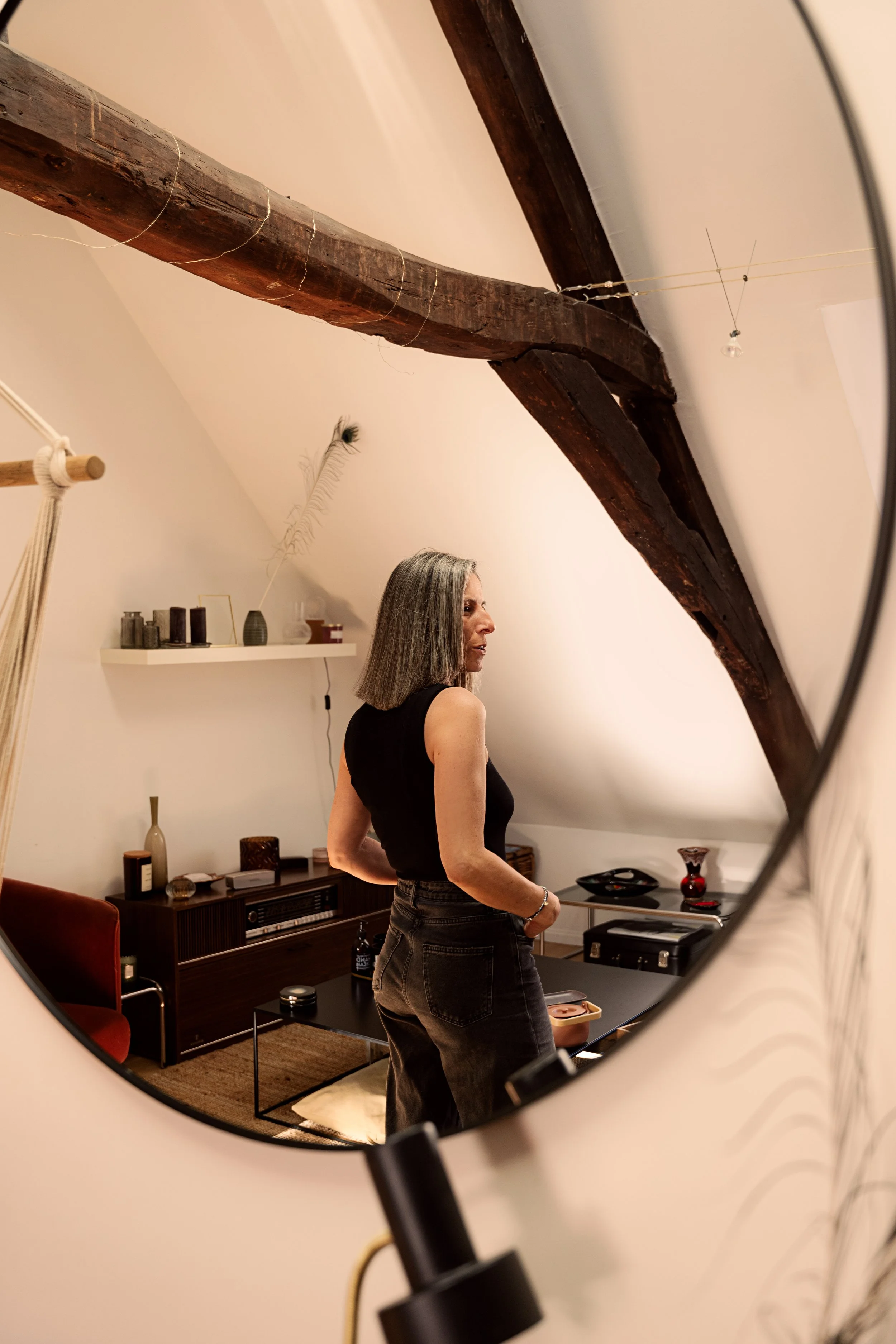 Reflet d'une femme aux cheveux gris dans un miroir rond, dans un salon moderne avec des éléments décoratifs, des meubles en bois foncé et une poutre en bois au plafond.