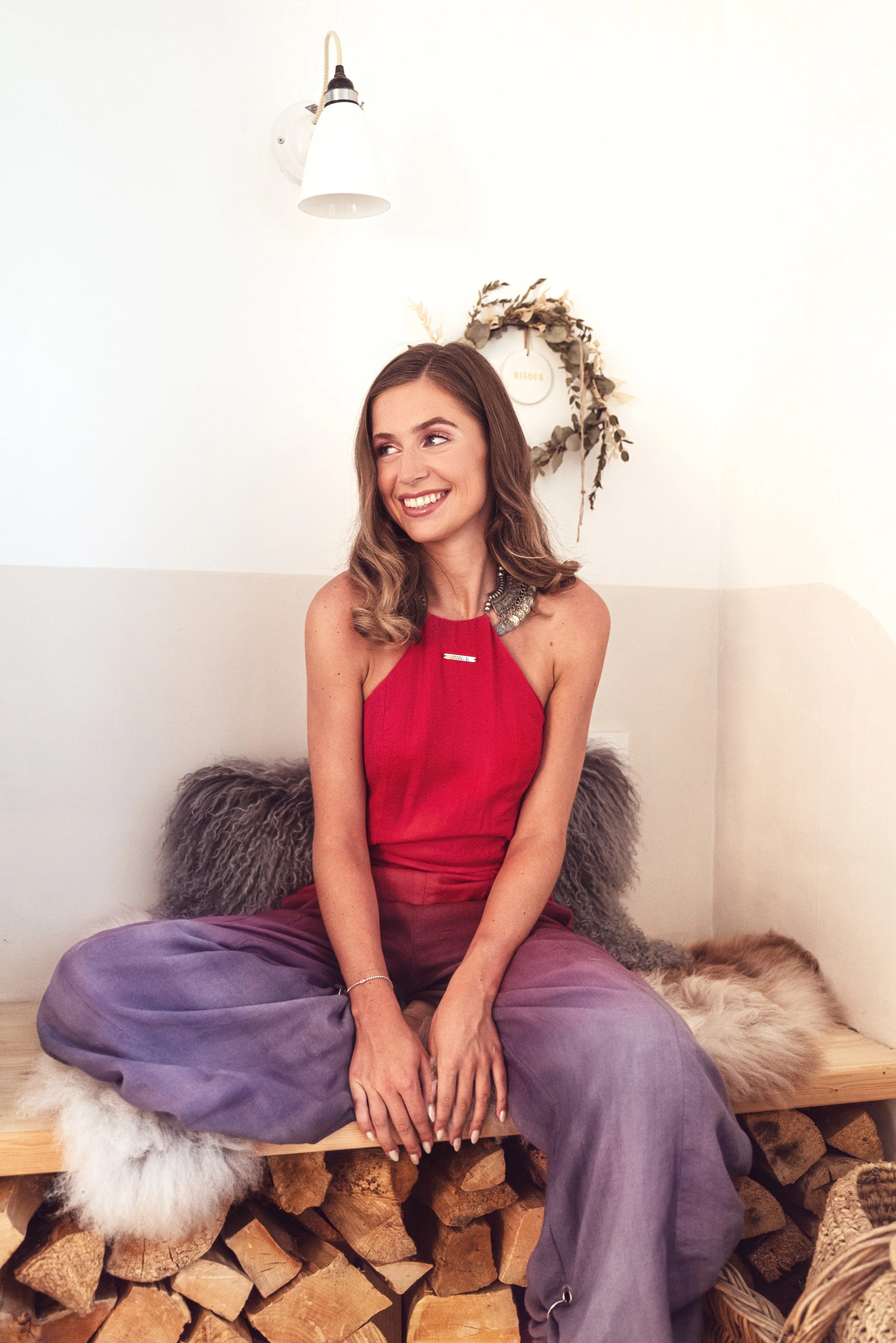 Une femme souriante assise sur une caisse en bois, portant un haut rouge et un pantalon violet, avec des coussins en fausse fourrure derrière elle, dans une pièce avec une décoration naturelle et minimaliste.