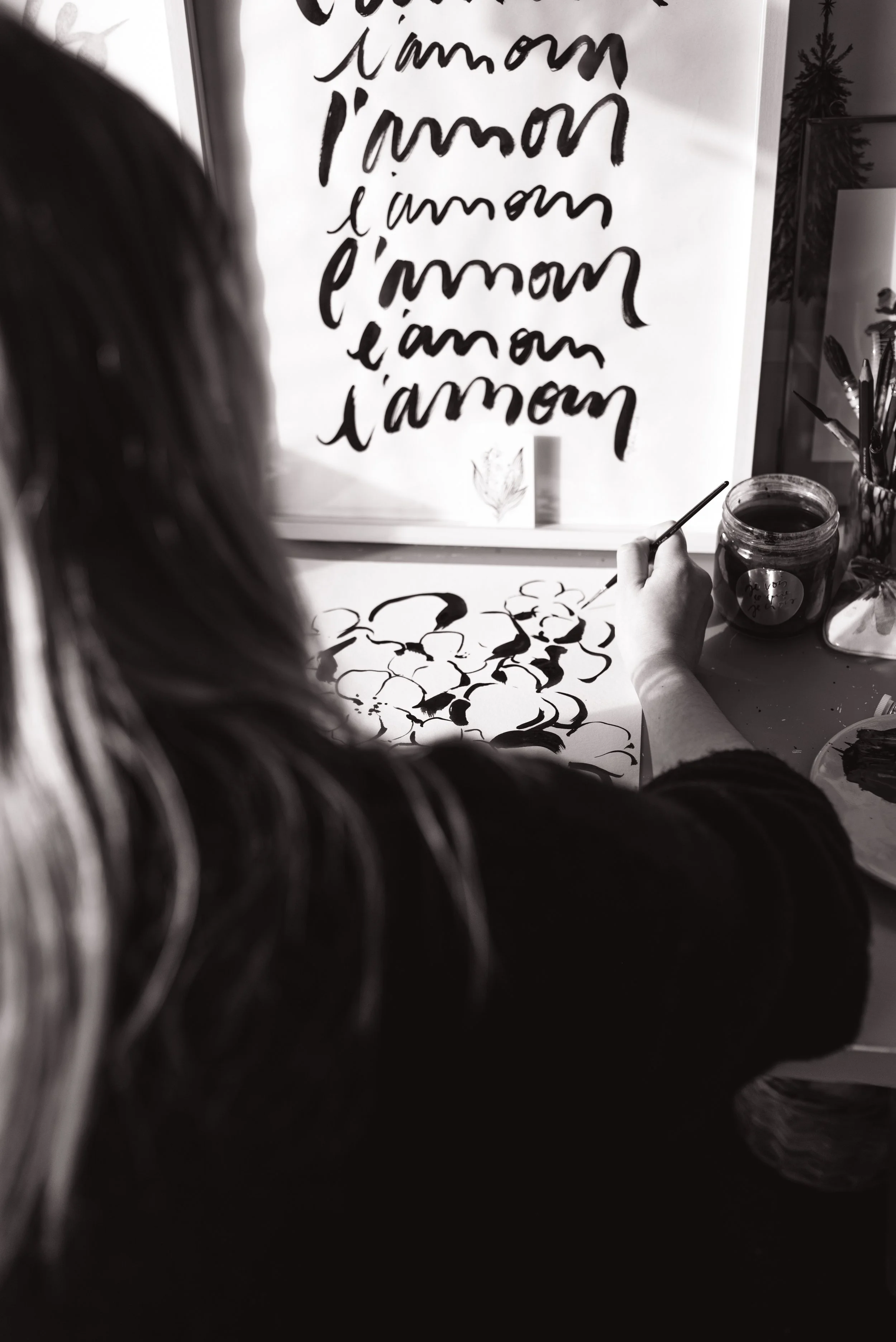 Une artiste peint de l'encre noire sur une feuille, avec un tableau en arrière-plan avec l'inscription 'l'amour' répétée en lettres cursives.