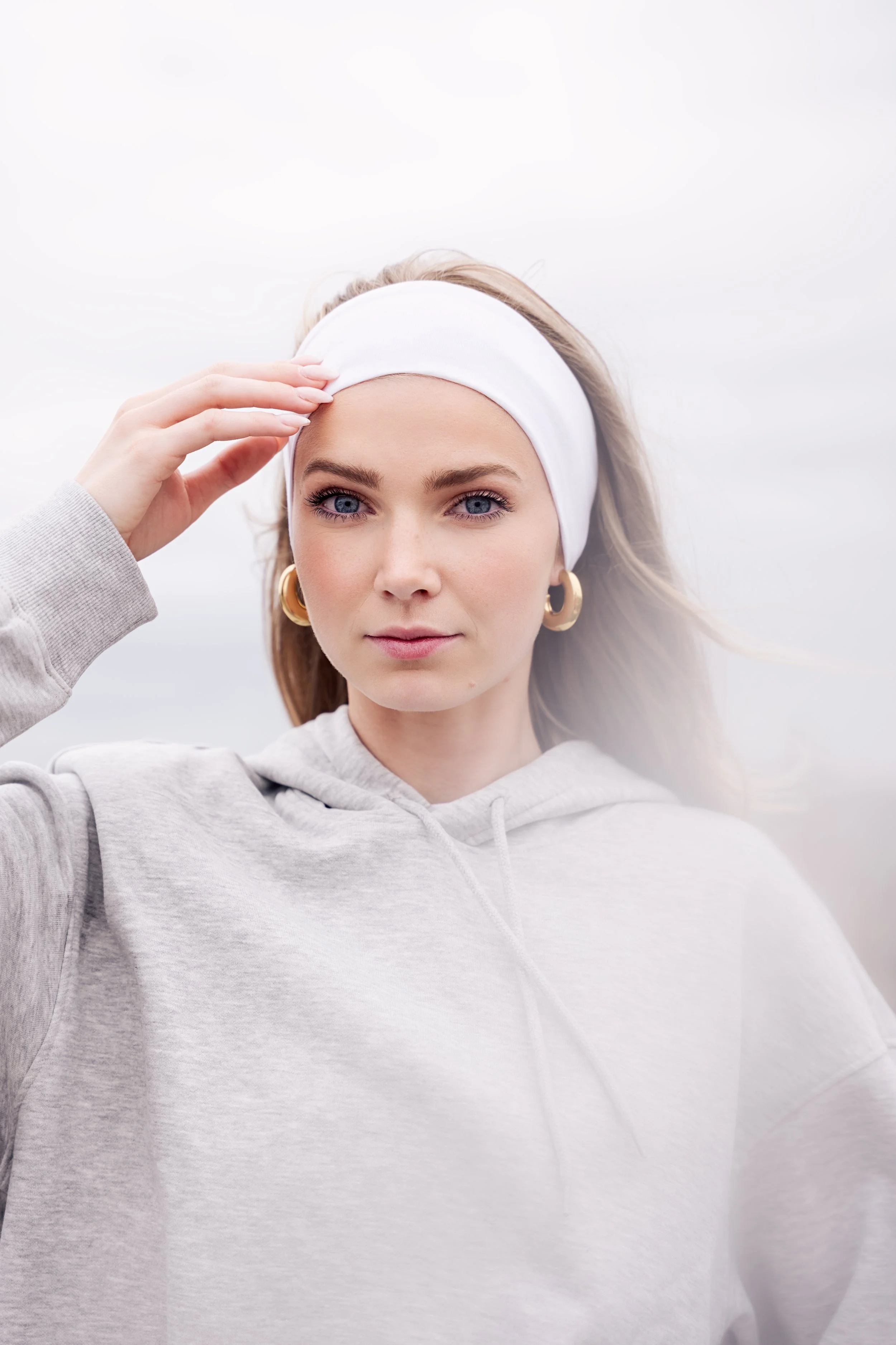 Photographe portrait Lille, Paris, Lyon. Jeune femme portant un bandeau blanc, sweat à capuche gris clair, boucles d'oreilles épaisses, regardant vers l'avant, arrière-plan nuageux.