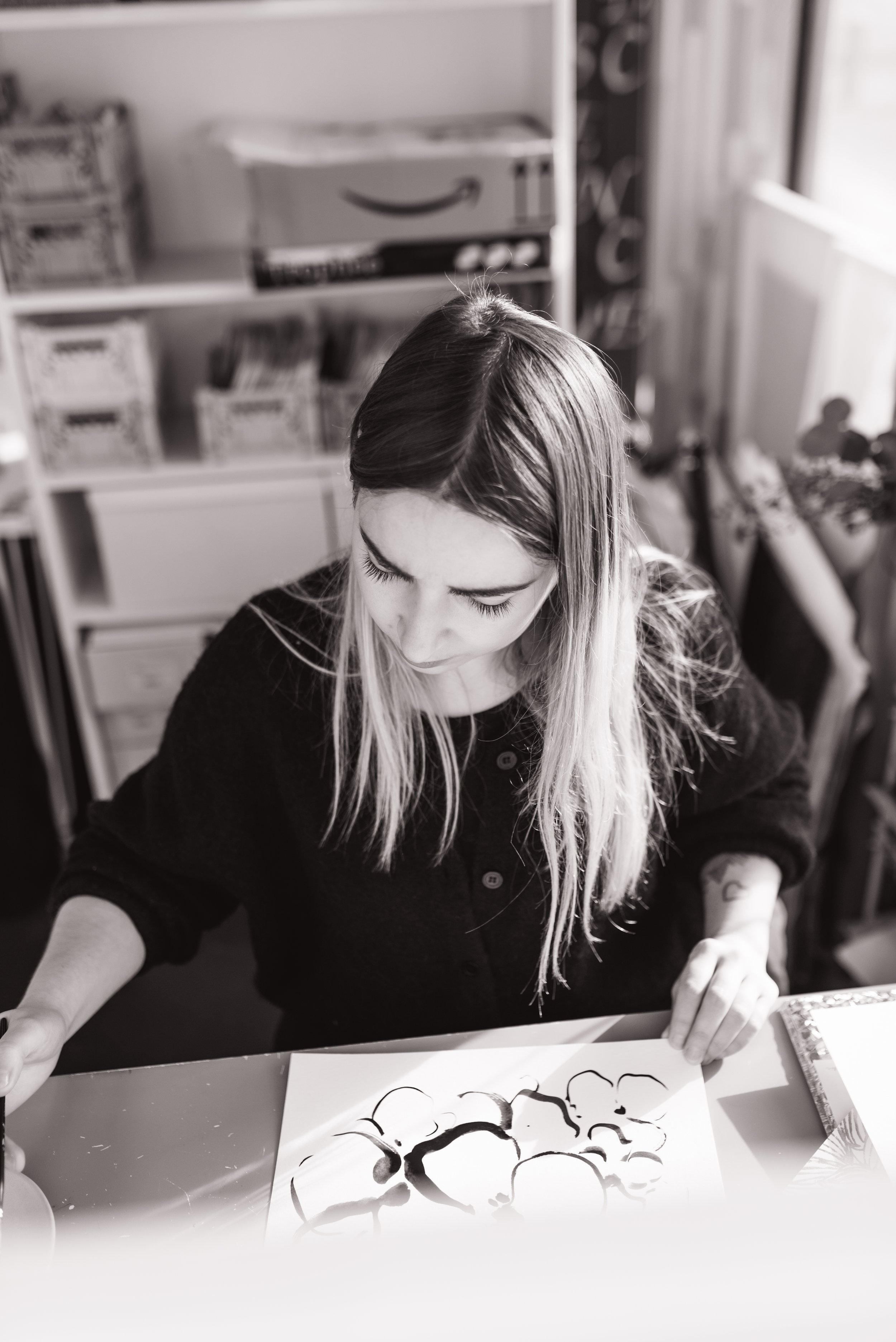 Reportage photo créateur pour la Presse. Jeune femme dessinant une œuvre abstraite à l'encre sur une feuille, vue de dessus, en intérieur, en noir et blanc.