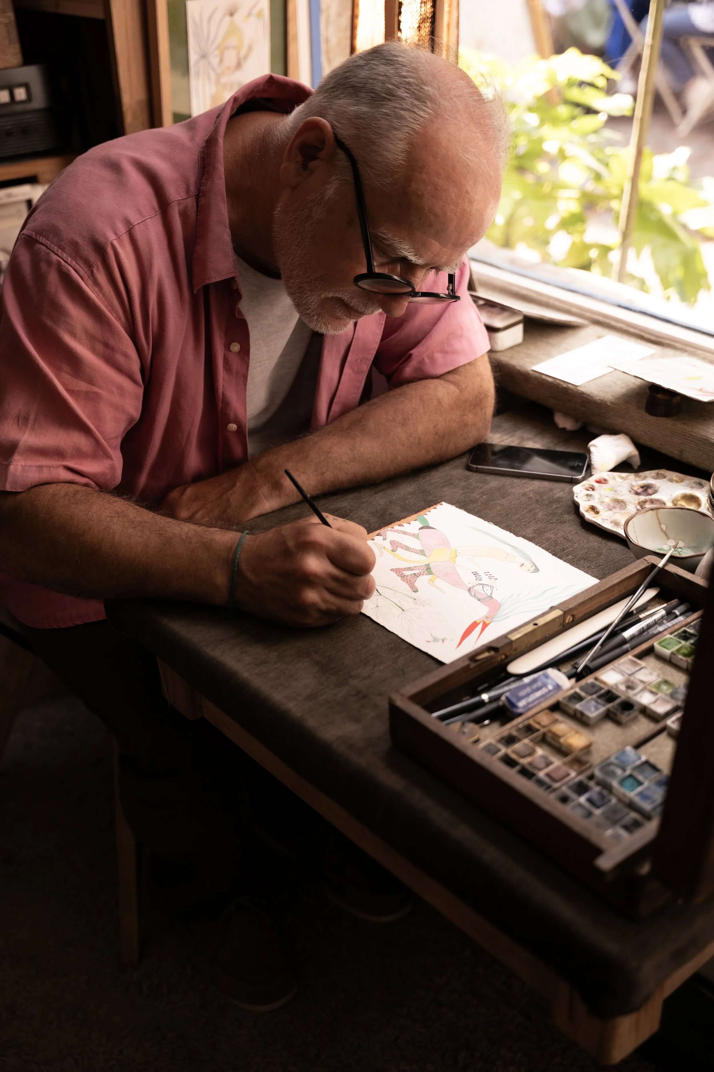 Un homme âgé, portant des lunettes et une chemise rose, assis à une table en train de peindre une illustration d'un animal fantastique coloré.
