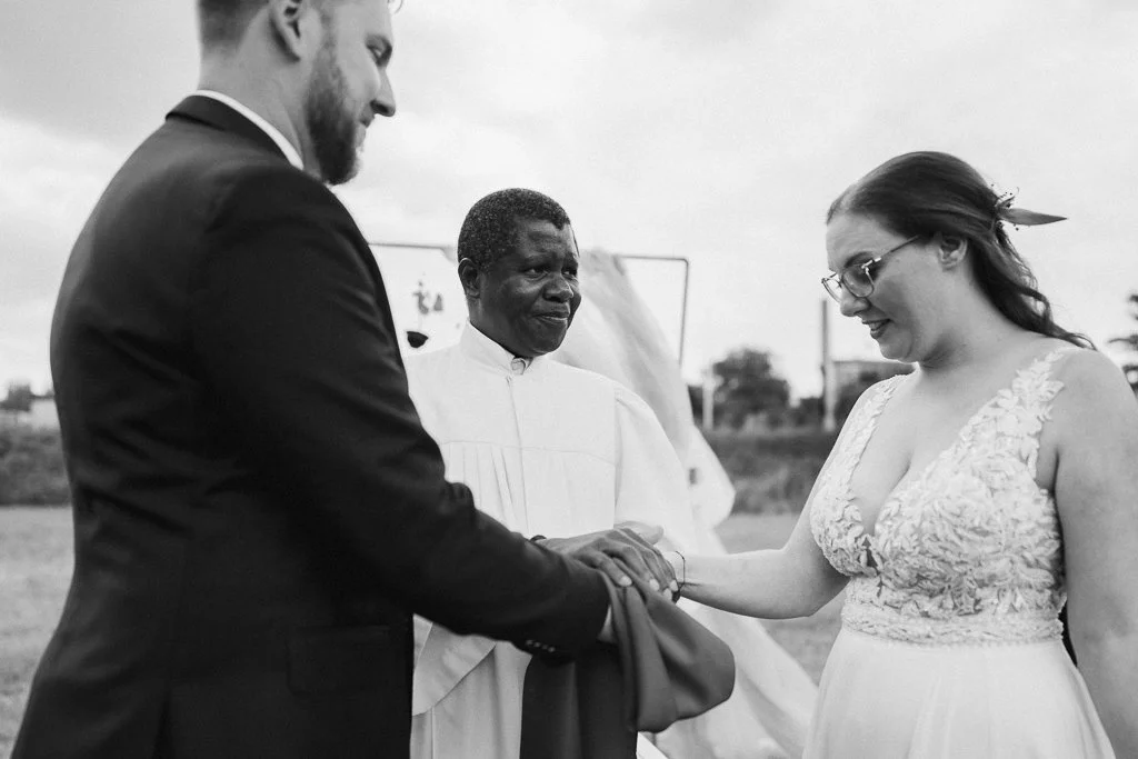 A wedding ceremony outdoors with three people: a bride in a lace dress and glasses, a groom in a suit, and an officiant in white robes, holding hands and exchanging vows.