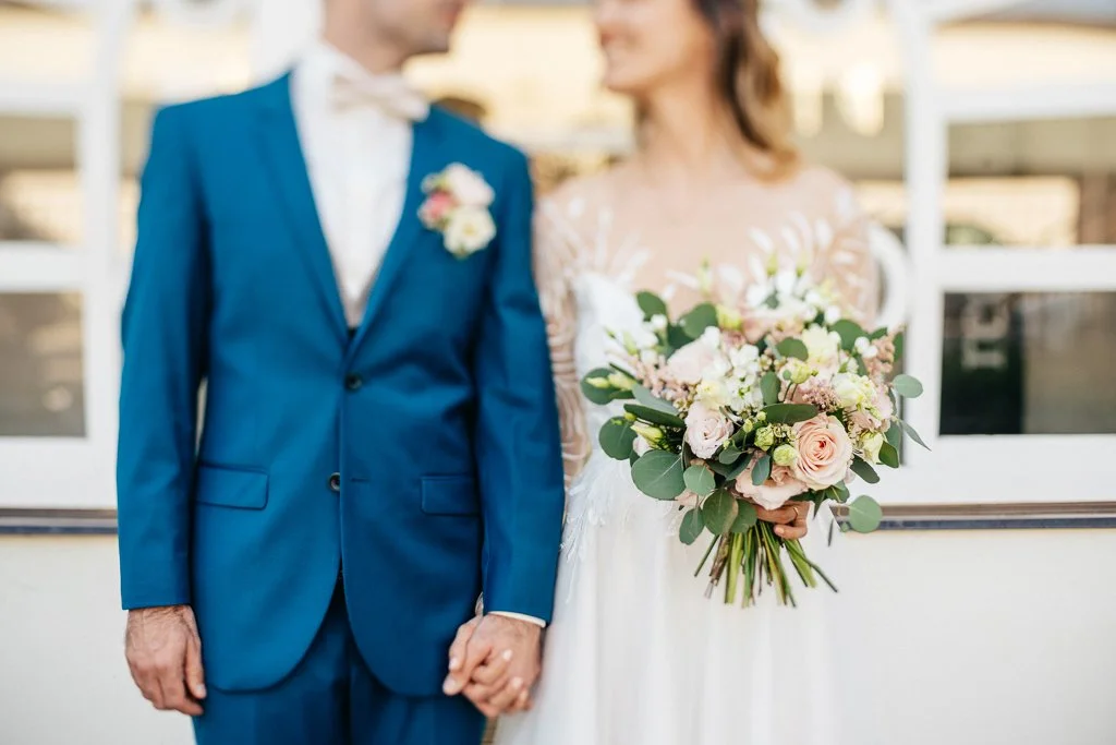 A bride holding a bouquet of pink and white flowers standing next to a groom in a blue suit at a wedding.