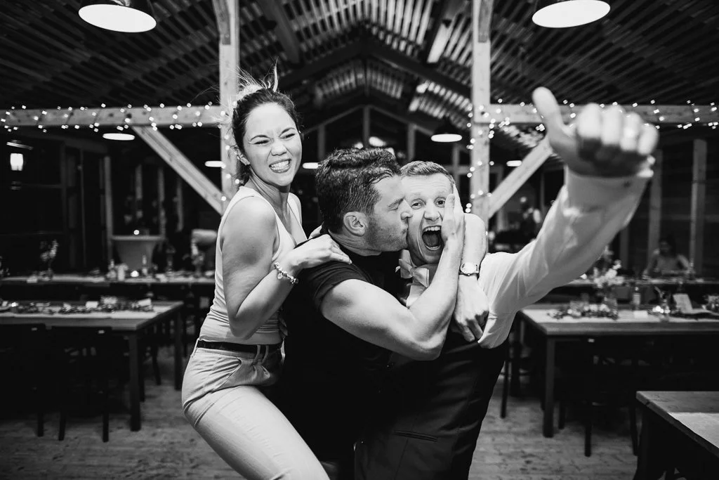 Group of three people at a party or celebration, laughing and having fun, indoors with string lights and decorated tables in the background.