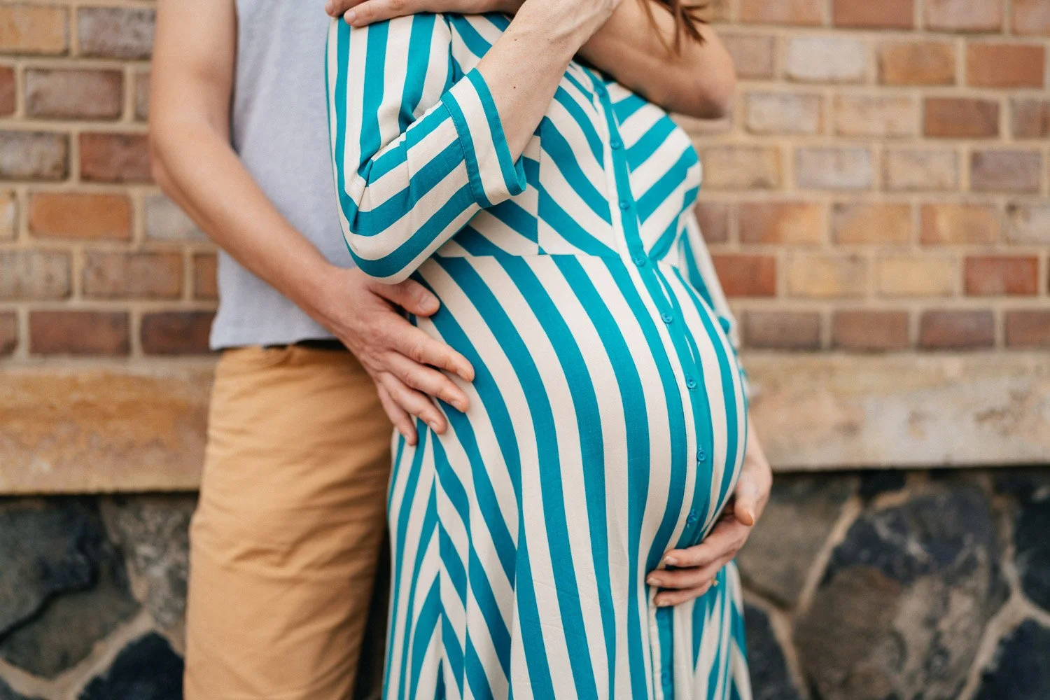 Detail rukou Andrey a Filipa na bříšku před cihlovou zdí.