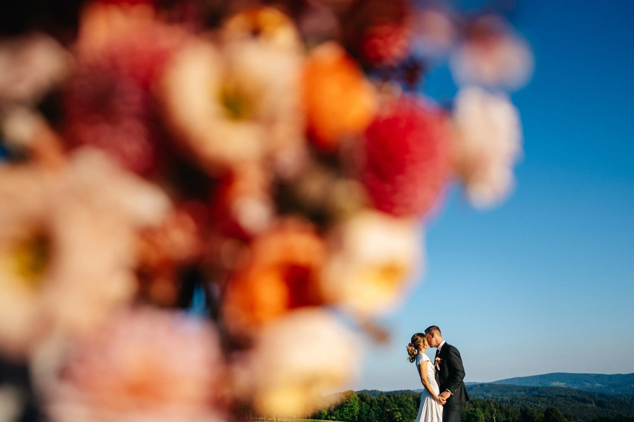 A blurred foreground of colorful berries with a clear background of a bride and groom holding hands, leaning in for a kiss, outdoors under a blue sky with hills in the distance.
