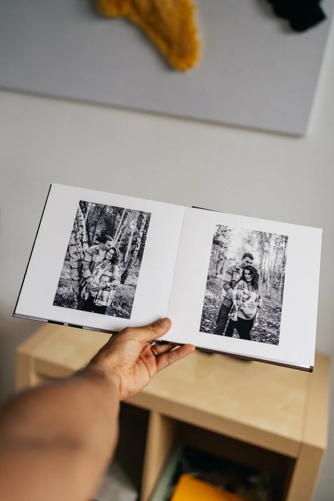Person holding an open photo album with two black and white pictures of a couple in a forest setting. The album is being held over a light wood table with books underneath.