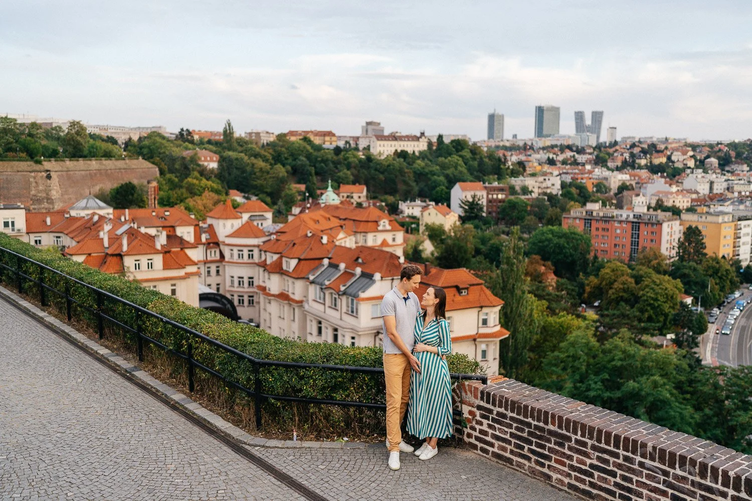 Pár u kamenného zábradlí s výhledem na pražské střechy a stromy.