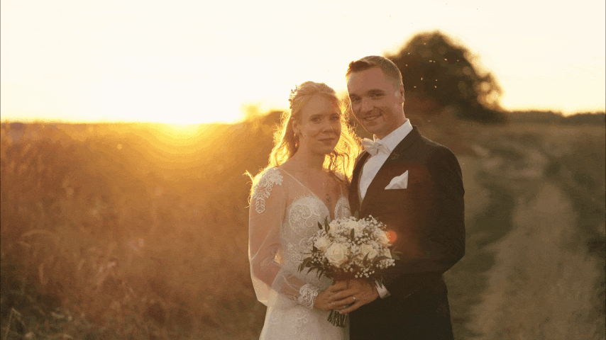 A newlywed couple posing outdoors at sunset, with the bride in a lace wedding gown holding a floral bouquet, and the groom in a tuxedo, smiling.