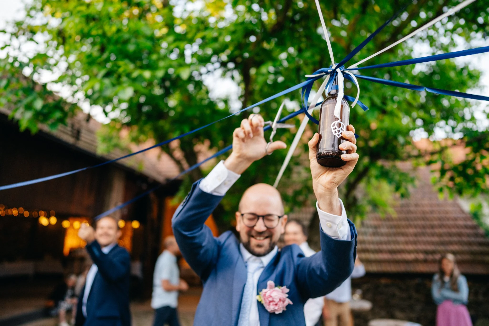 Man in blue suit at outdoor event holding beer bottle with blue and white ribbons.