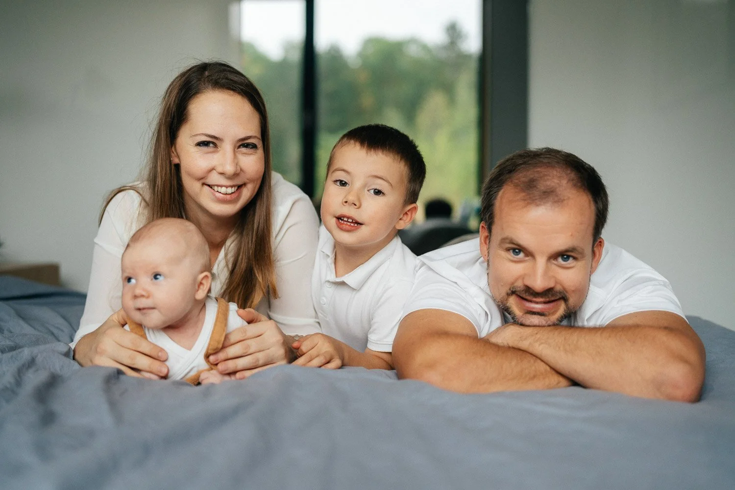 Rodina leží společně na posteli a dívá se do fotoaparátu.