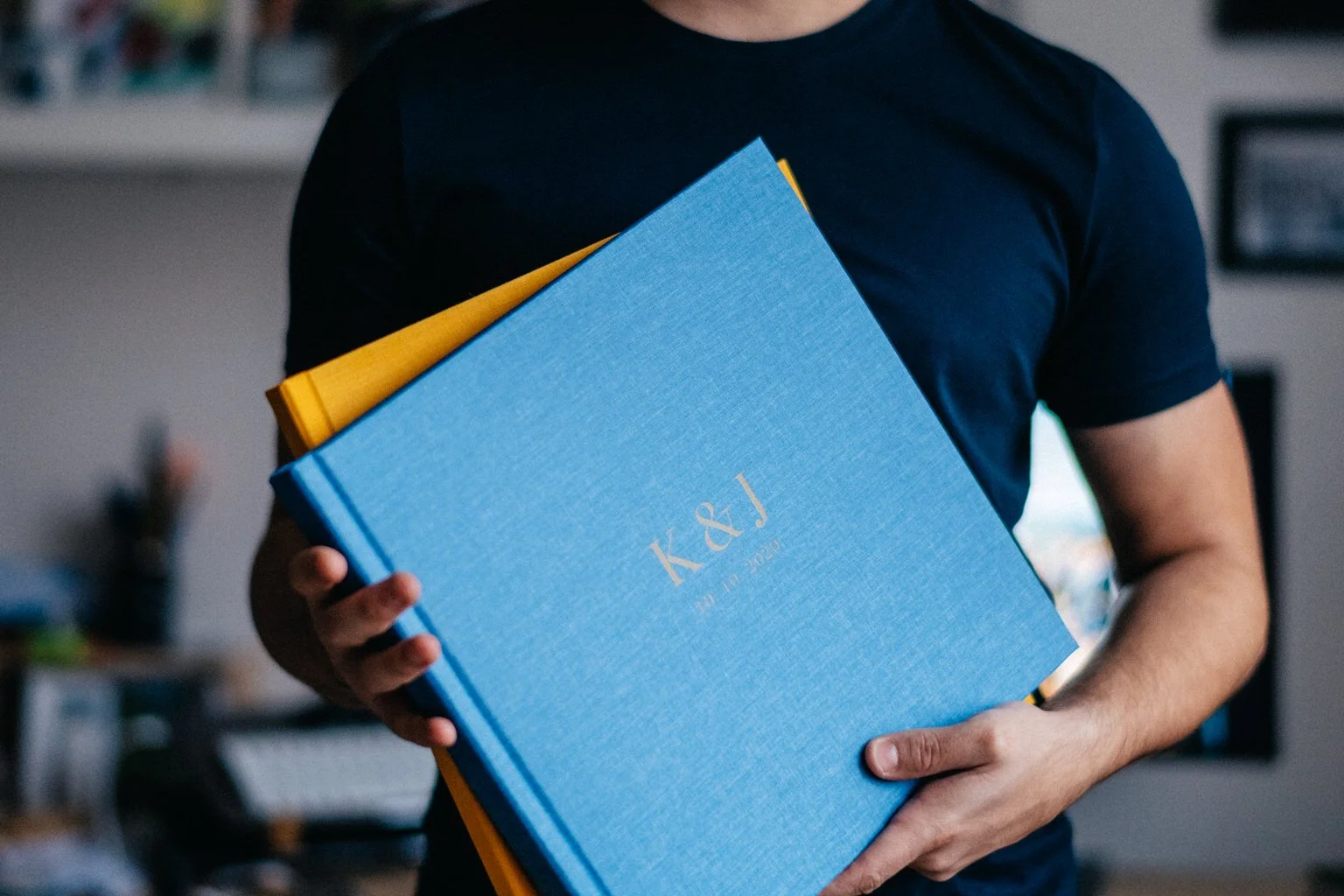Person holding a blue photo album with initials K&J, partially covering a yellow album, in an indoor setting.