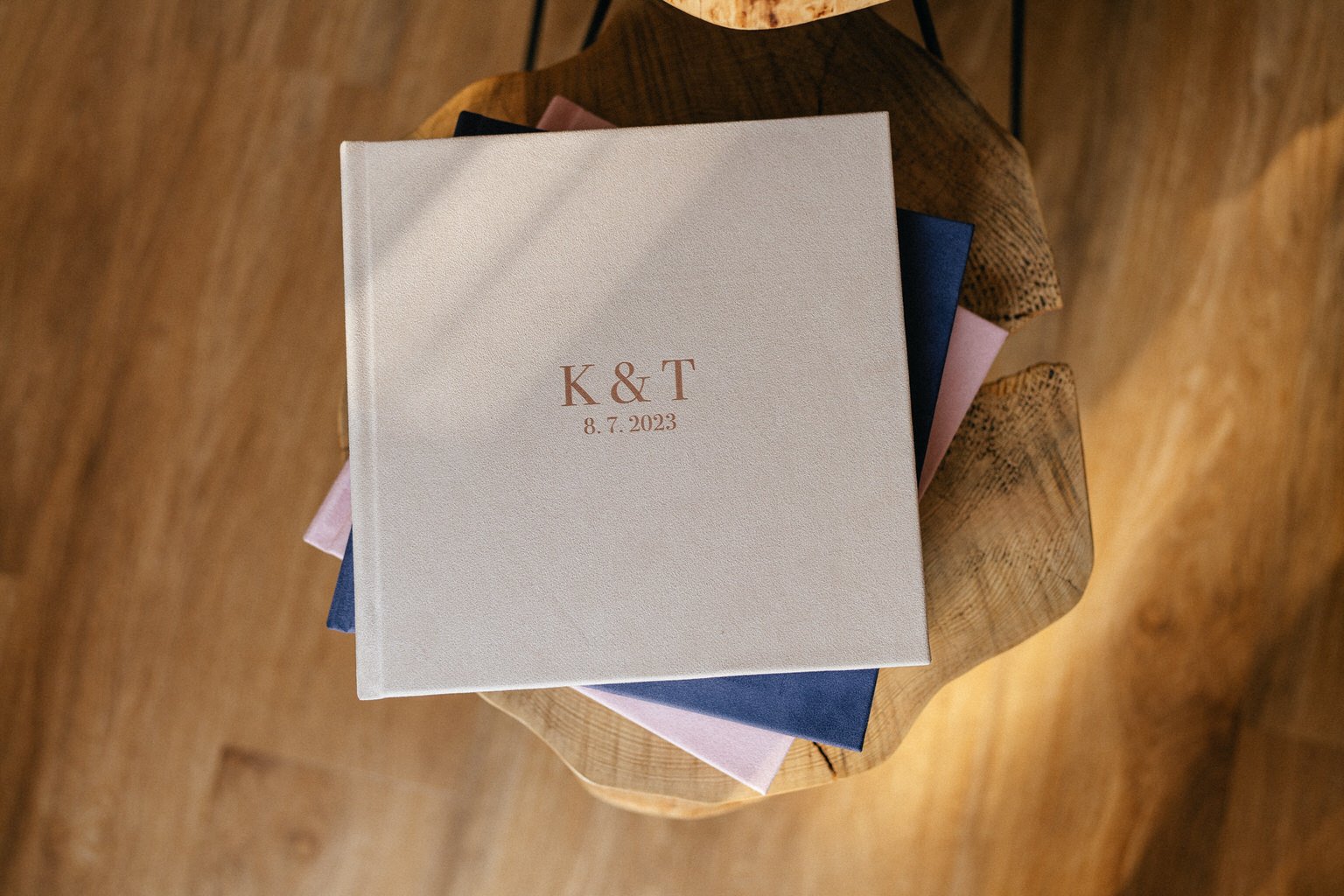 A beige book with "K & T 8.7.2023" embossed on the cover, placed on a wooden surface with a pink and blue book underneath.