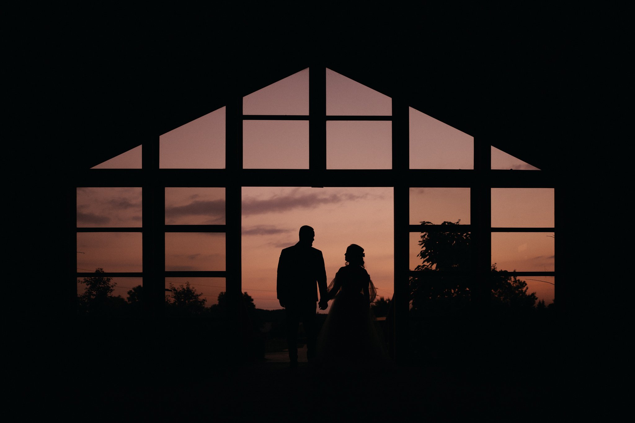 Silhouette of a couple holding hands inside a house with large windows during a sunset.