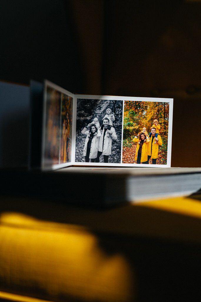 An open photo album on a table, displaying black and white and color family photos with people wearing coats in a park setting with autumn leaves.