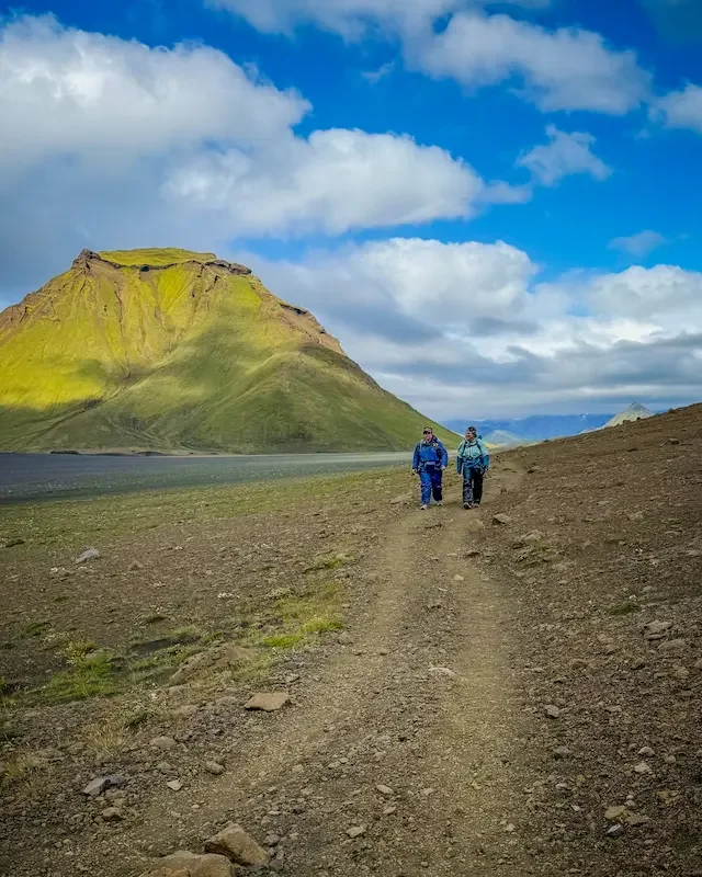 hikers on trail walking beside water on laugavegur trek and green hill beside