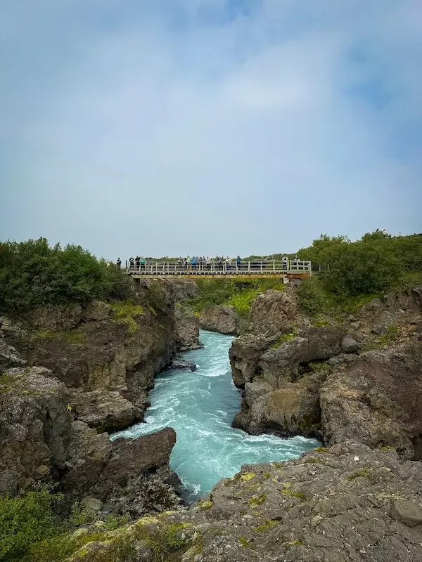 blue river through rocks and group of people standing on bridge above